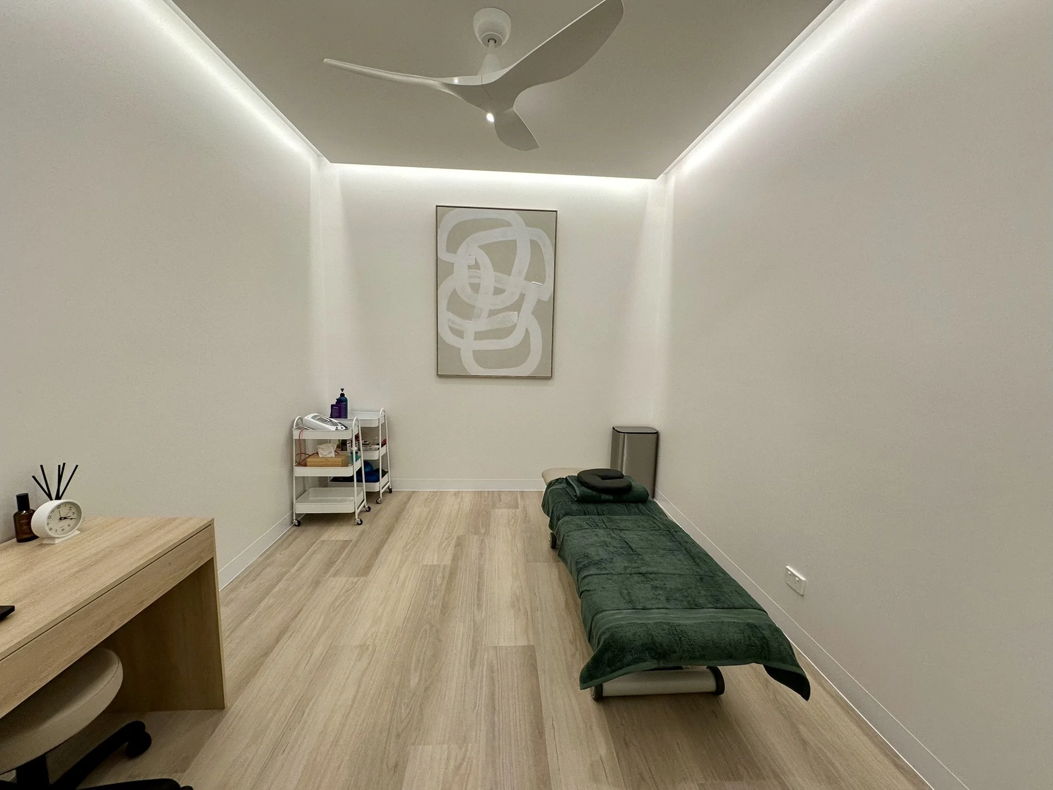 Room with white walls, ceiling fan, framed abstract artwork, green massage or treatment table, small white shelf with bottles and supplies, clock, and wooden desk with decorative items.