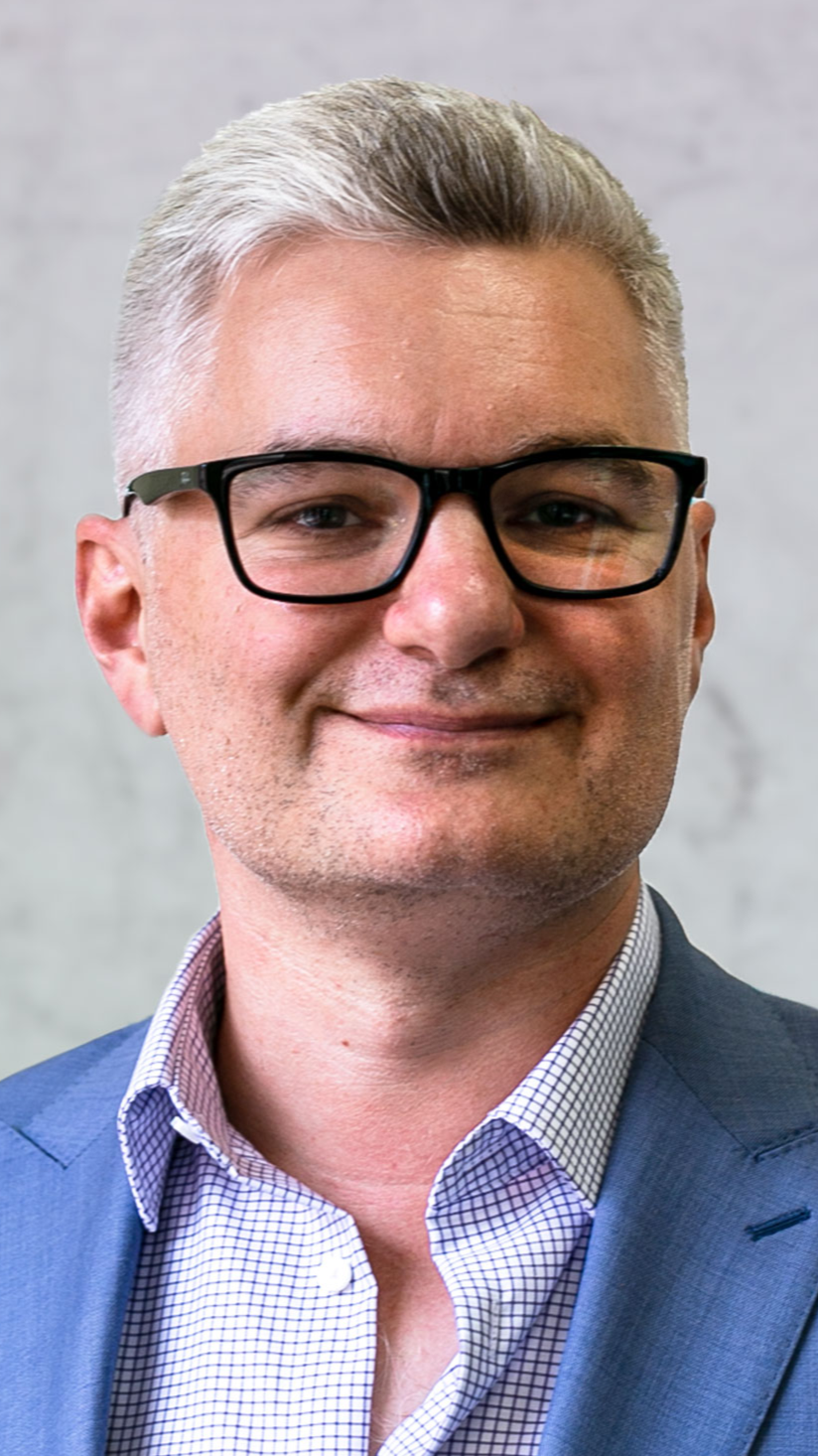 Portrait of a man with short gray hair and black glasses, wearing a blue suit and a checkered shirt, smiling in front of a plain gray wall.