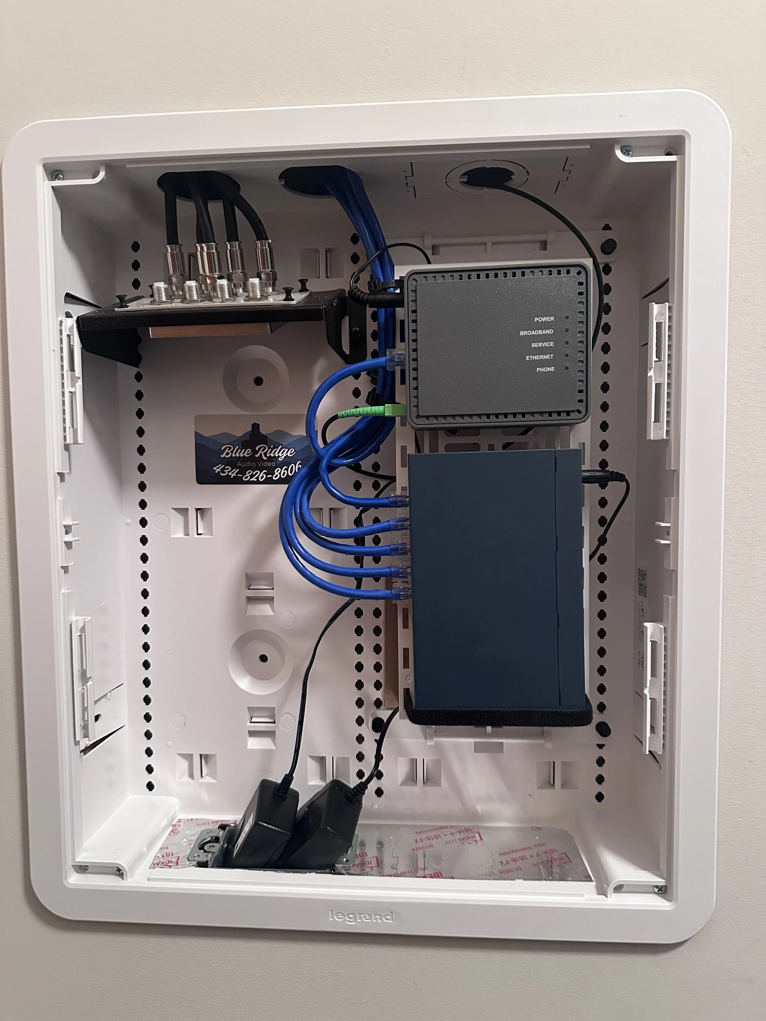 Open fiber optic internet connection box with blue cables and black power adapters inside a white wall-mounted enclosure.