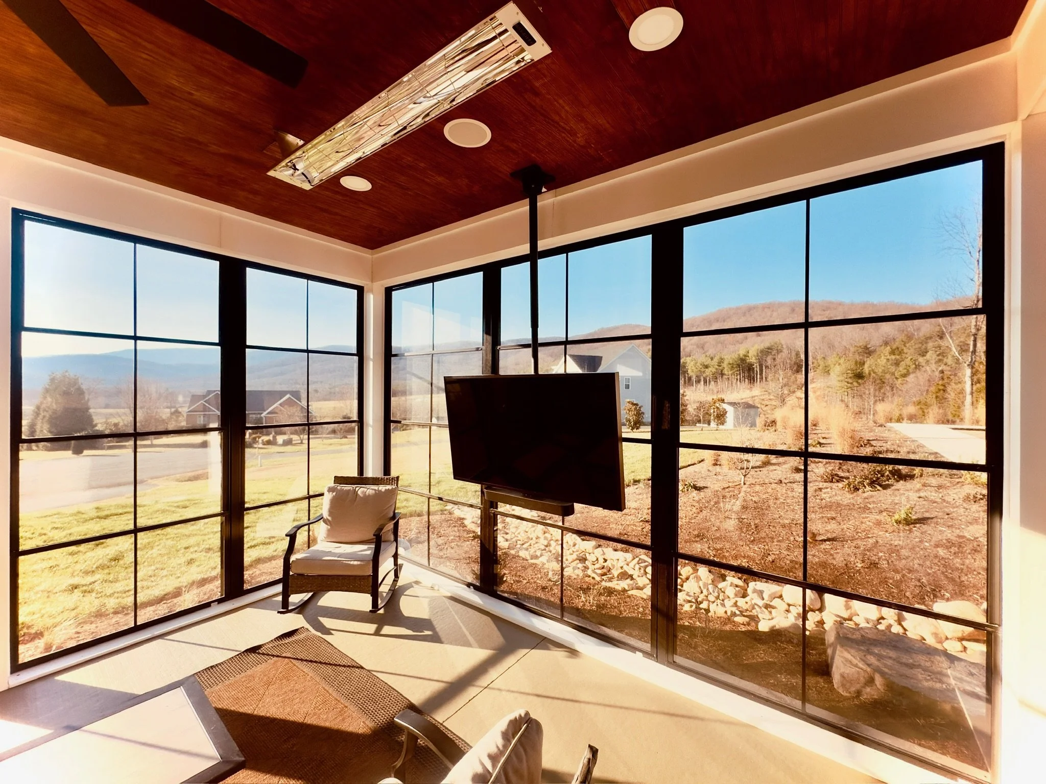 Interior view of a sunroom or enclosed porch with large floor-to-ceiling windows showing a view of a grassy yard, trees, and distant hills on a sunny day. There is a white chair with a pillow and a outdoor TV Mounted from the ceiling.