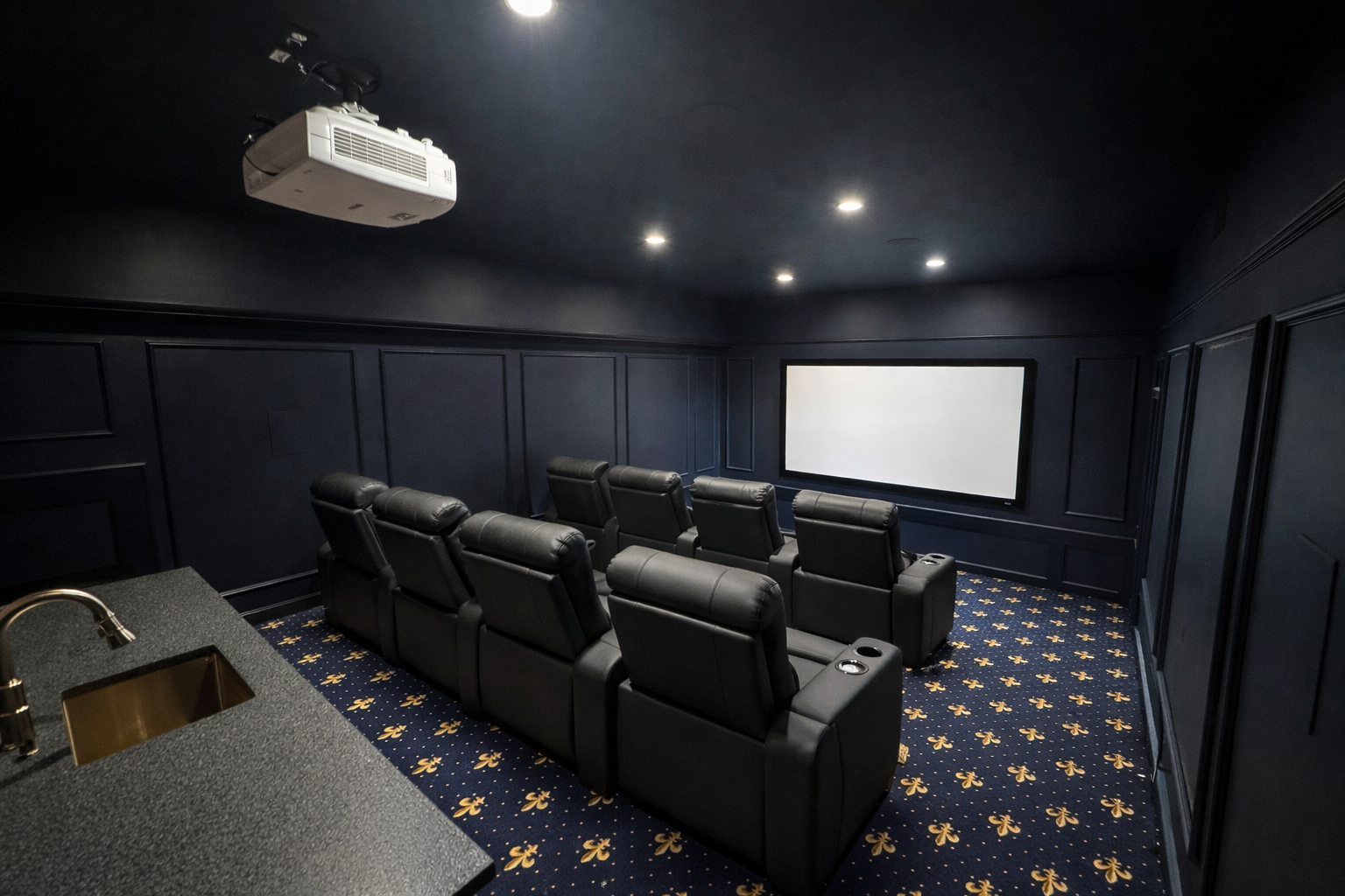 A home theater room with black walls, ceiling, and leather recliner seats facing a large white screen. The room is dimly lit by ceiling lights, and there is a projector mounted on the ceiling. A bar area with a sink and countertop is visible in the foreground.