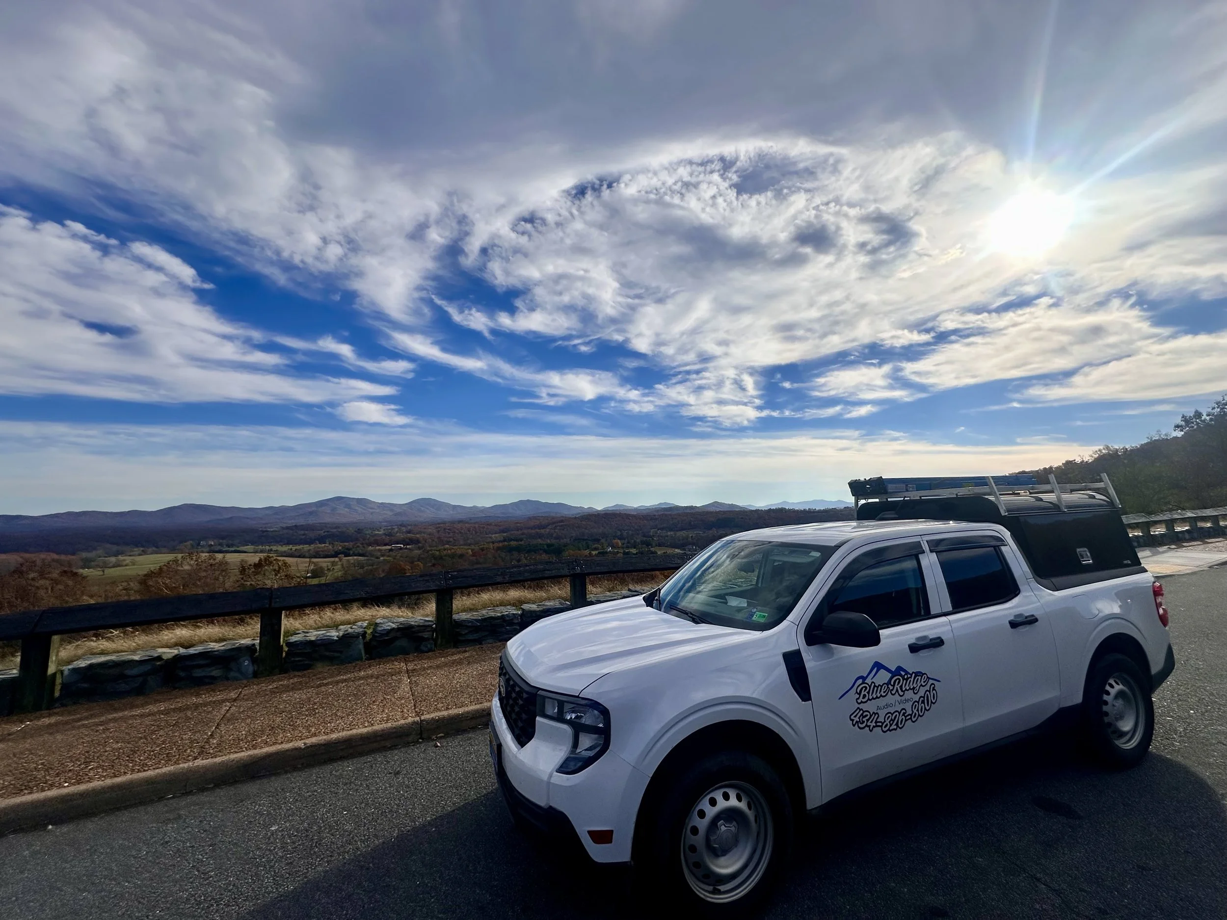 A white Ford Maverick with a logo for Blue Ridge Audio Video and a phone number parked next to a guardrail overlooking a vast landscape of mountains and fields under a partly cloudy sky with the sun shining.