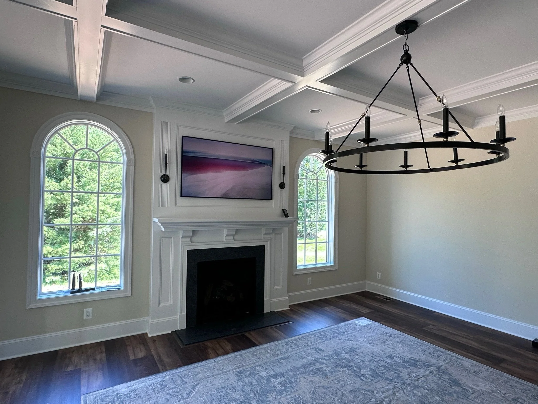 Living room with large arched windows, a wall-mounted Samsung Frame television, a fireplace, and a chandelier hanging from a coffered ceiling.