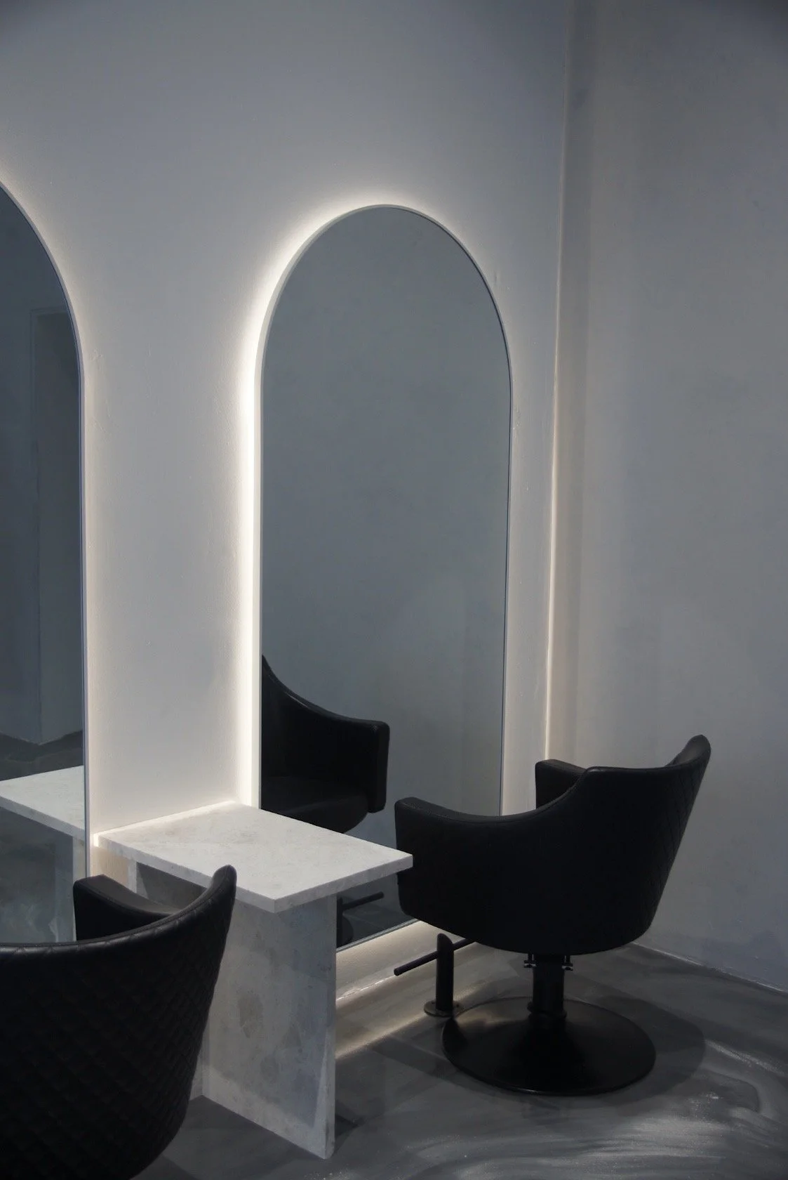 A modern salon or beauty studio setup with a large mirror framed by white LED lighting, a white marble table, and black salon chairs.