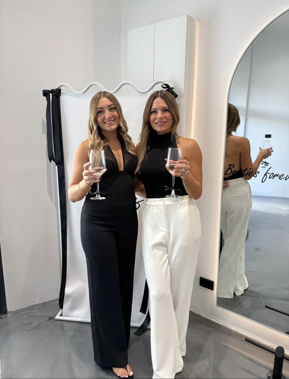 Two women standing together, smiling, holding wine glasses, dressed in black and white, in a room with a mirror that has the words "Betties forever" written on it.