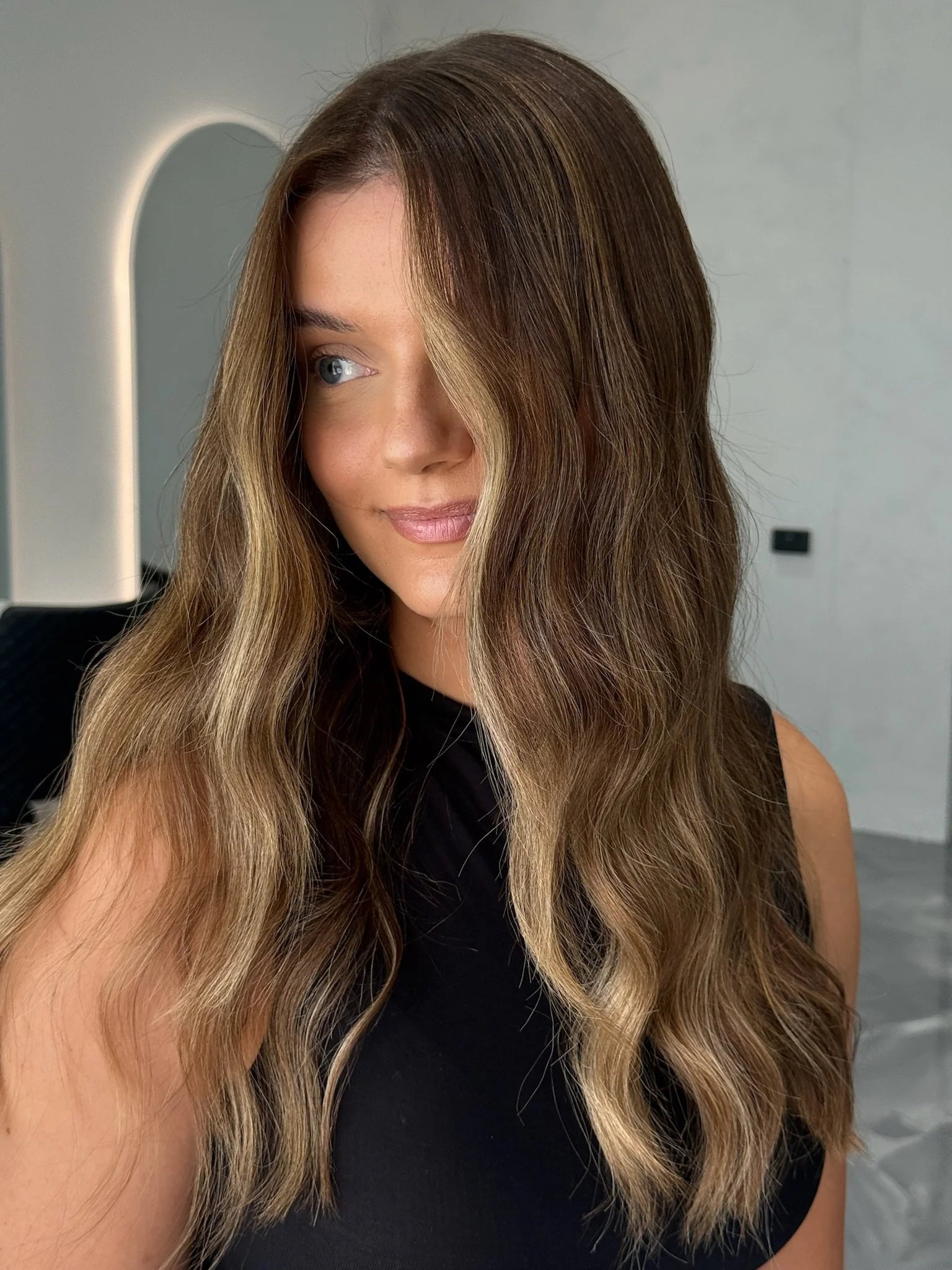 A woman with long, wavy hair that is dark brown with blonde highlights, wearing a black sleeveless top, looking off to the side with a slight smile, standing in a room with a light gray wall and a mirror in the background.