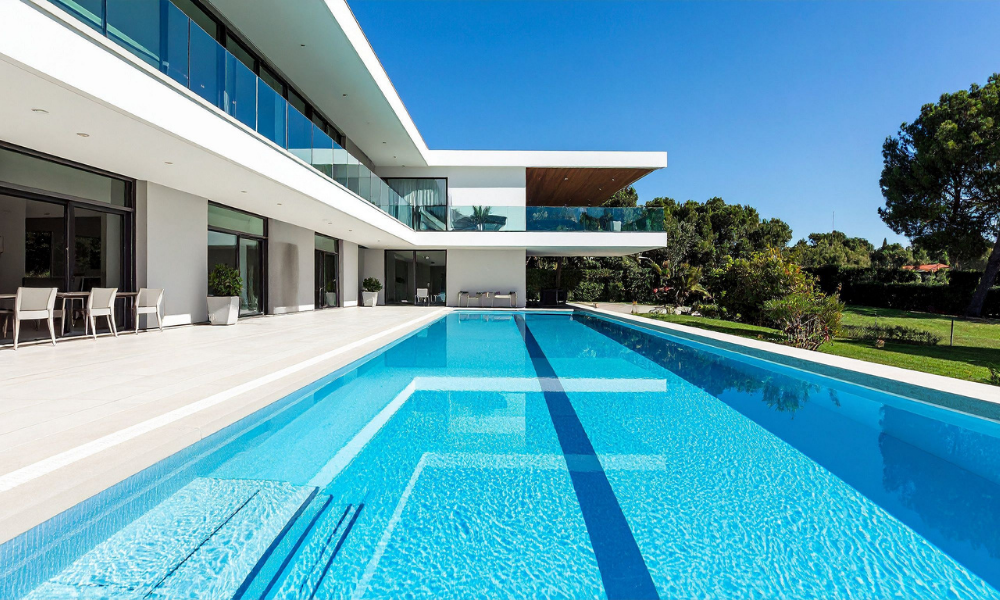 Modern white house with large outdoor swimming pool, glass balcony, and patio furniture, surrounded by green trees and a clear blue sky.