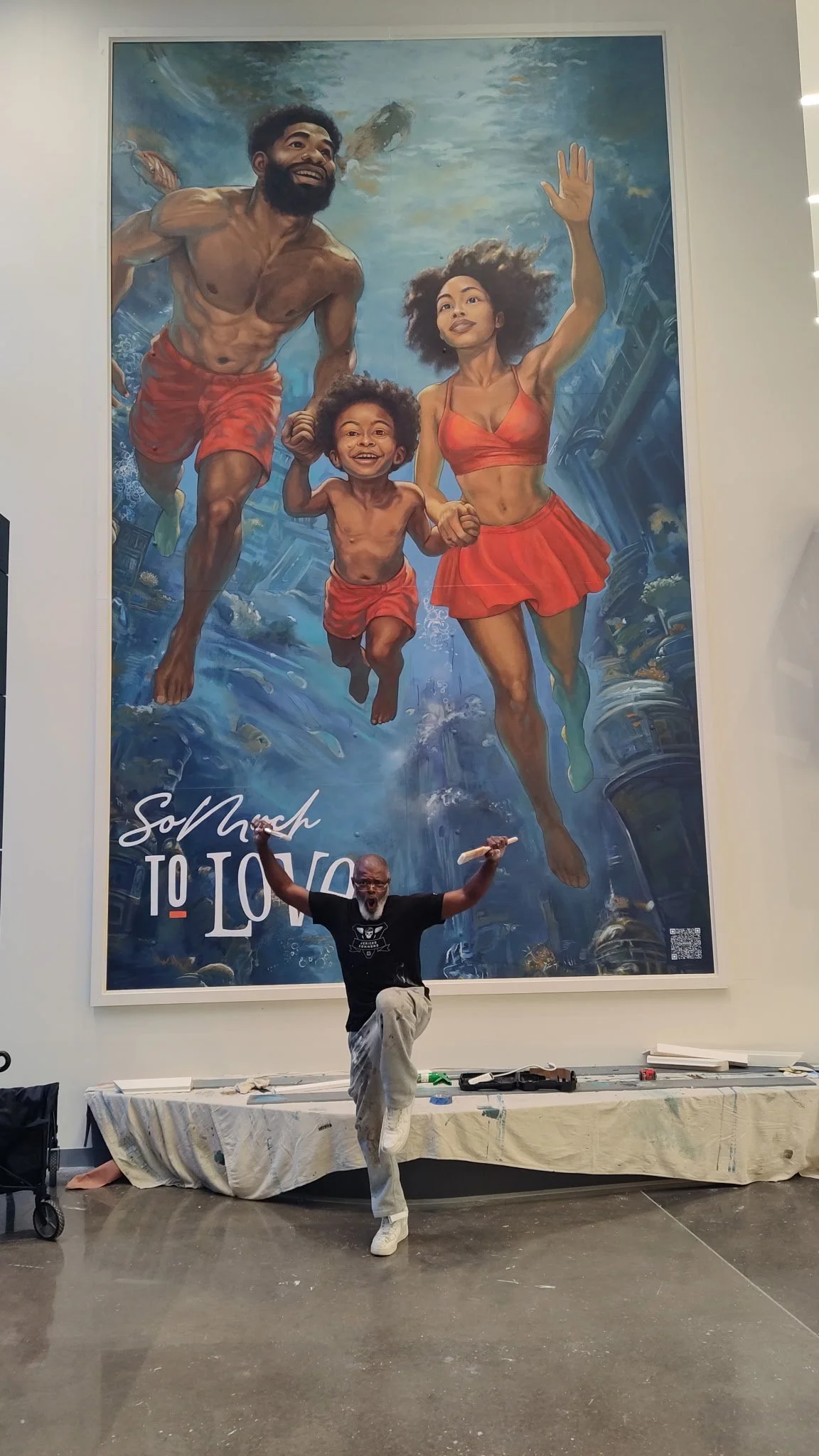 A man happily posing in front of a large colorful mural of a family running hand in hand. The mural depicts a man, woman, and child in bright red clothing, with a blue sky and ocean background. The man in the photo is holding a paintbrush.