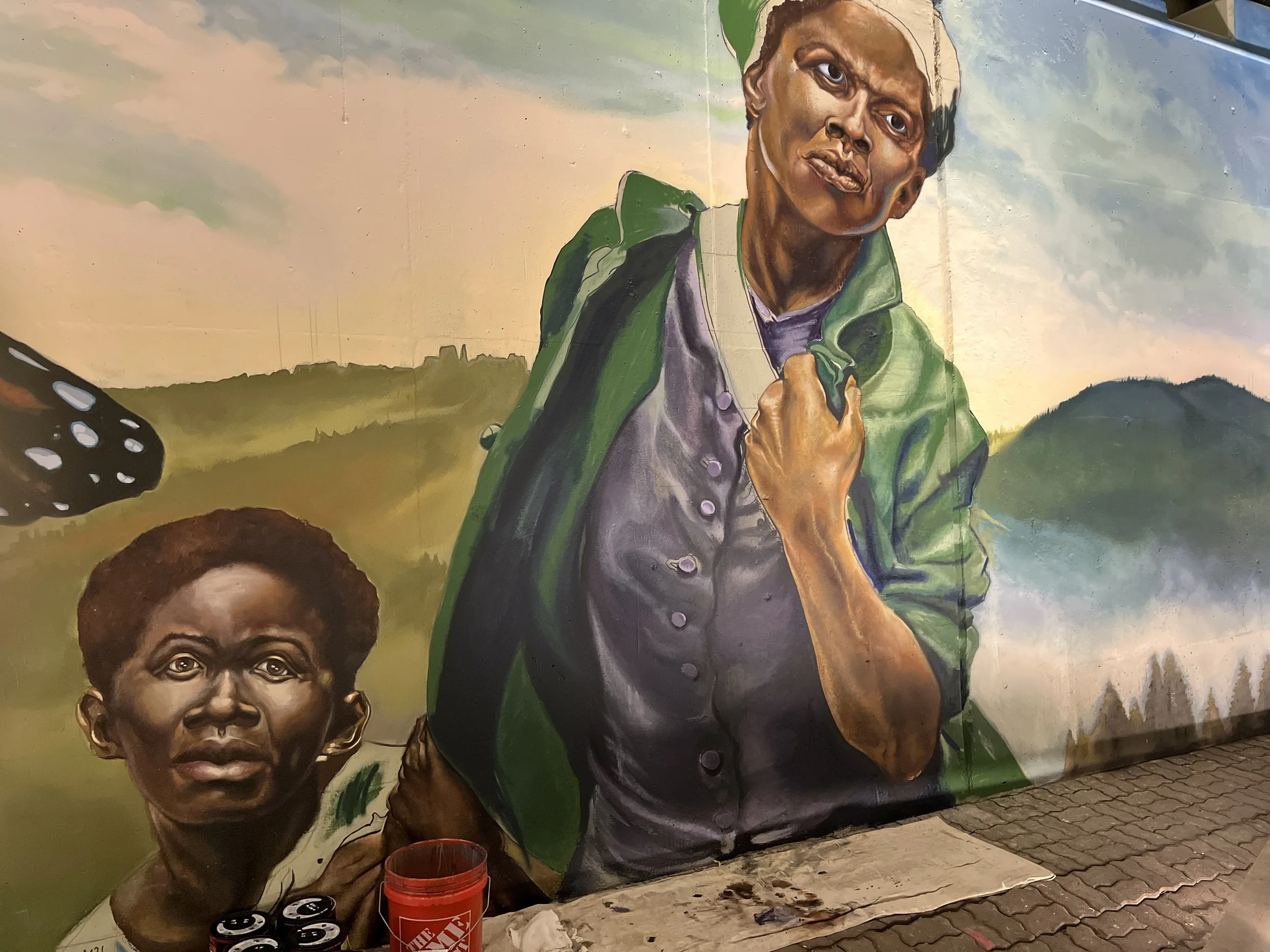 Mural painting of two women outdoors. One woman with short hair looks directly ahead, and the other woman with light hair is holding a strap of her green jacket and looking slightly upward. The background features a scenic landscape with hills and a 