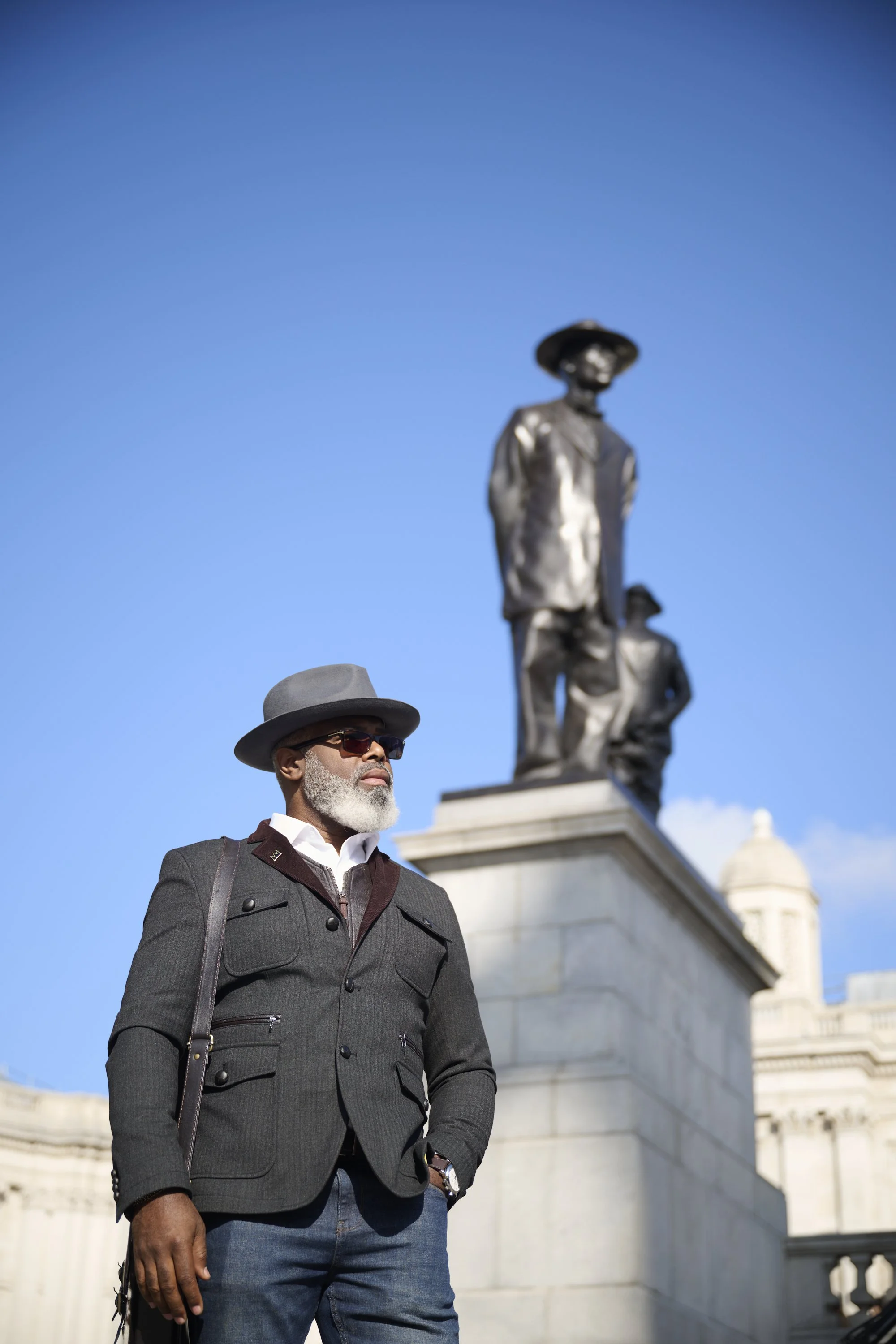A stylish older man with gray beard and sunglasses wearing a gray hat and jacket stands outdoors near a large stone statue of two soldiers against a clear blue sky.