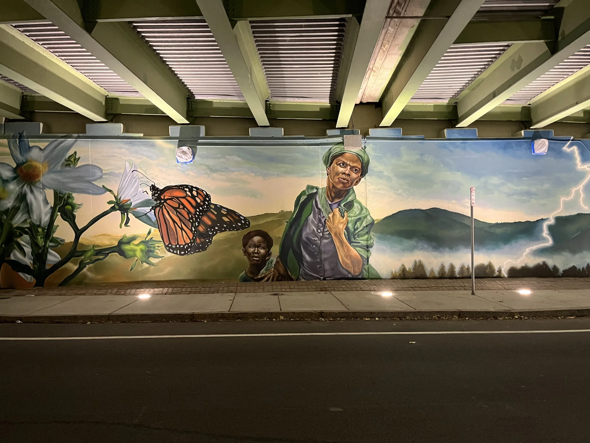 A large mural painted on a wall under a bridge, depicting a woman and a child, with butterflies, flowers, mountains, and lightning in the background.