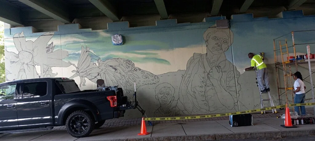 Artists work on a large mural under a bridge, with a black pickup truck parked in front and scaffolding on the right side.