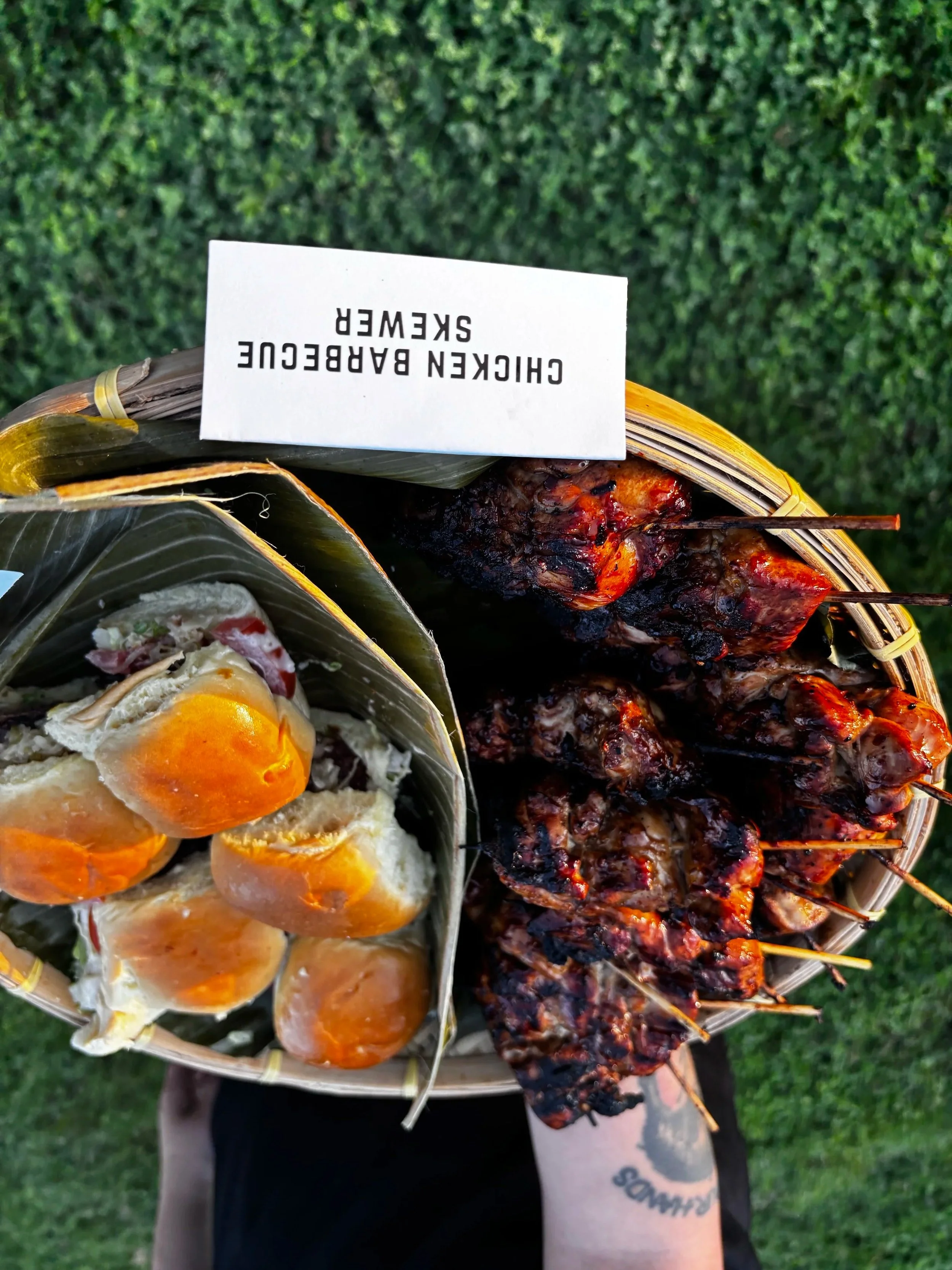 Skewers of grilled meat and a bamboo container with wrapped food, label reading 'Chicken Barbecue Skewer', outdoor grassy background.