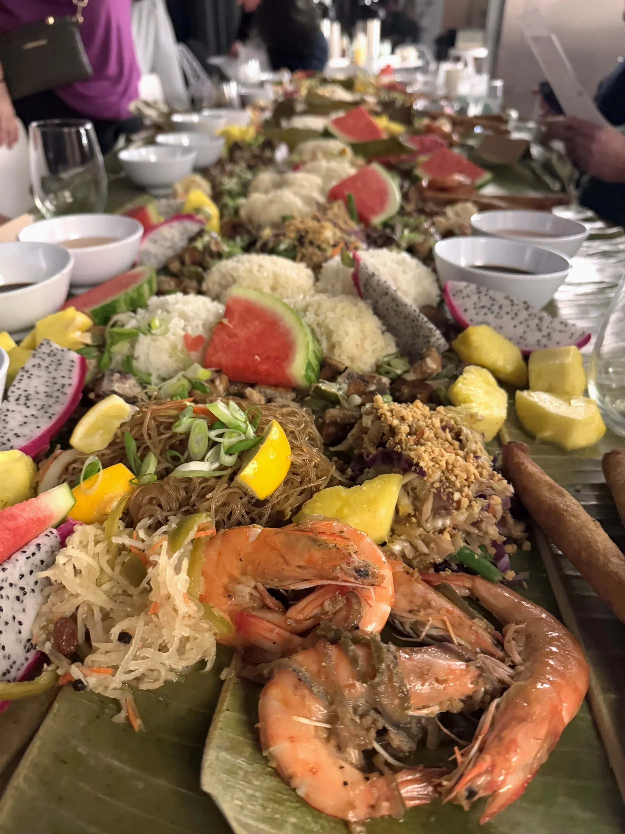 A large spread of various Asian dishes, including shrimp, noodles, fruits like dragon fruit, watermelon, pineapple, and dragon fruit, along with small bowls of sauces, on a long table.