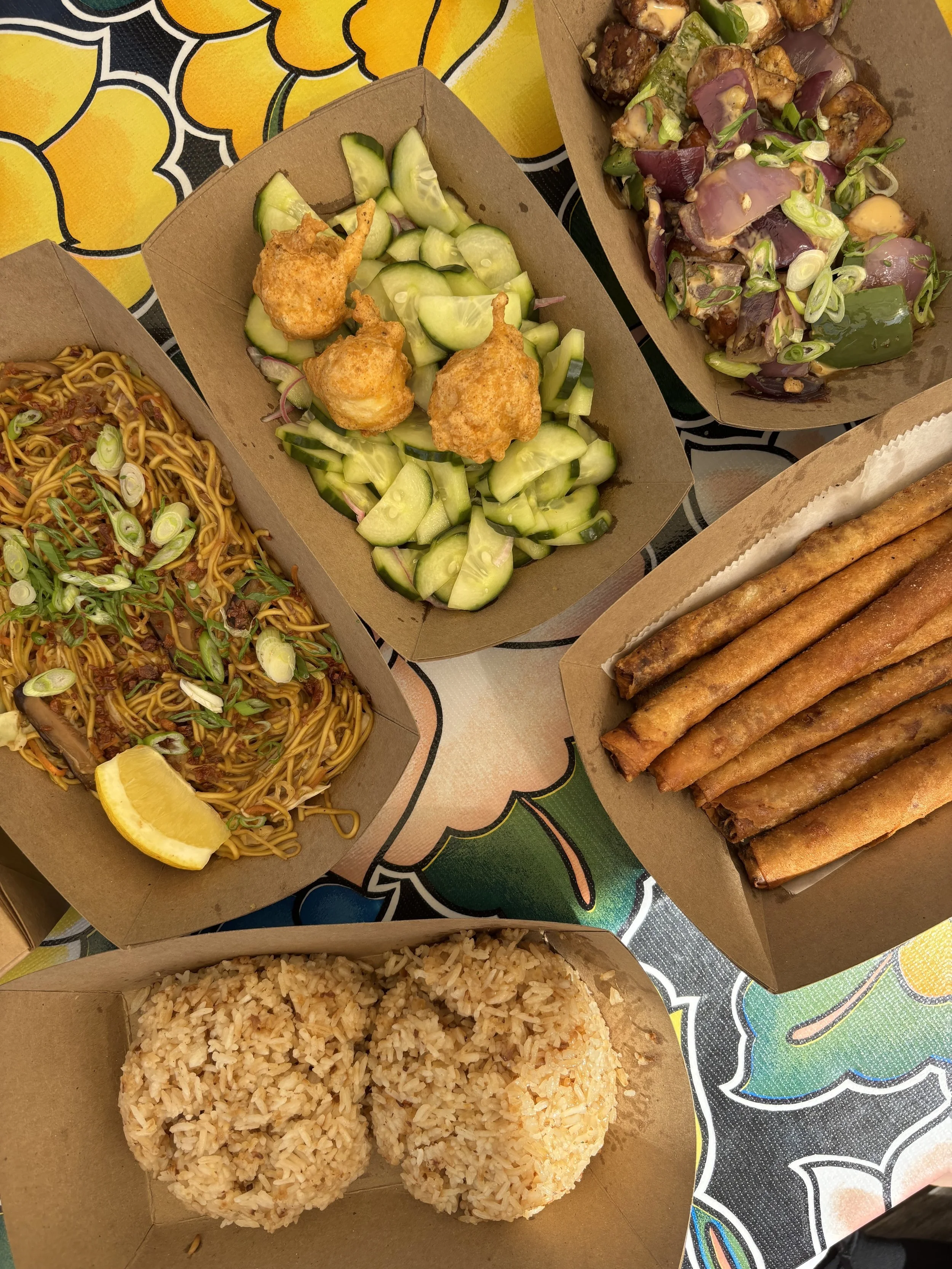 A variety of takeout food including fried rice, ramen noodles with lemon wedge, cucumber salad with fried shrimp, stakes topped with sauce and scallions, spring rolls, and grilled meat with vegetables, arranged in cardboard containers on a colorful patterned table.