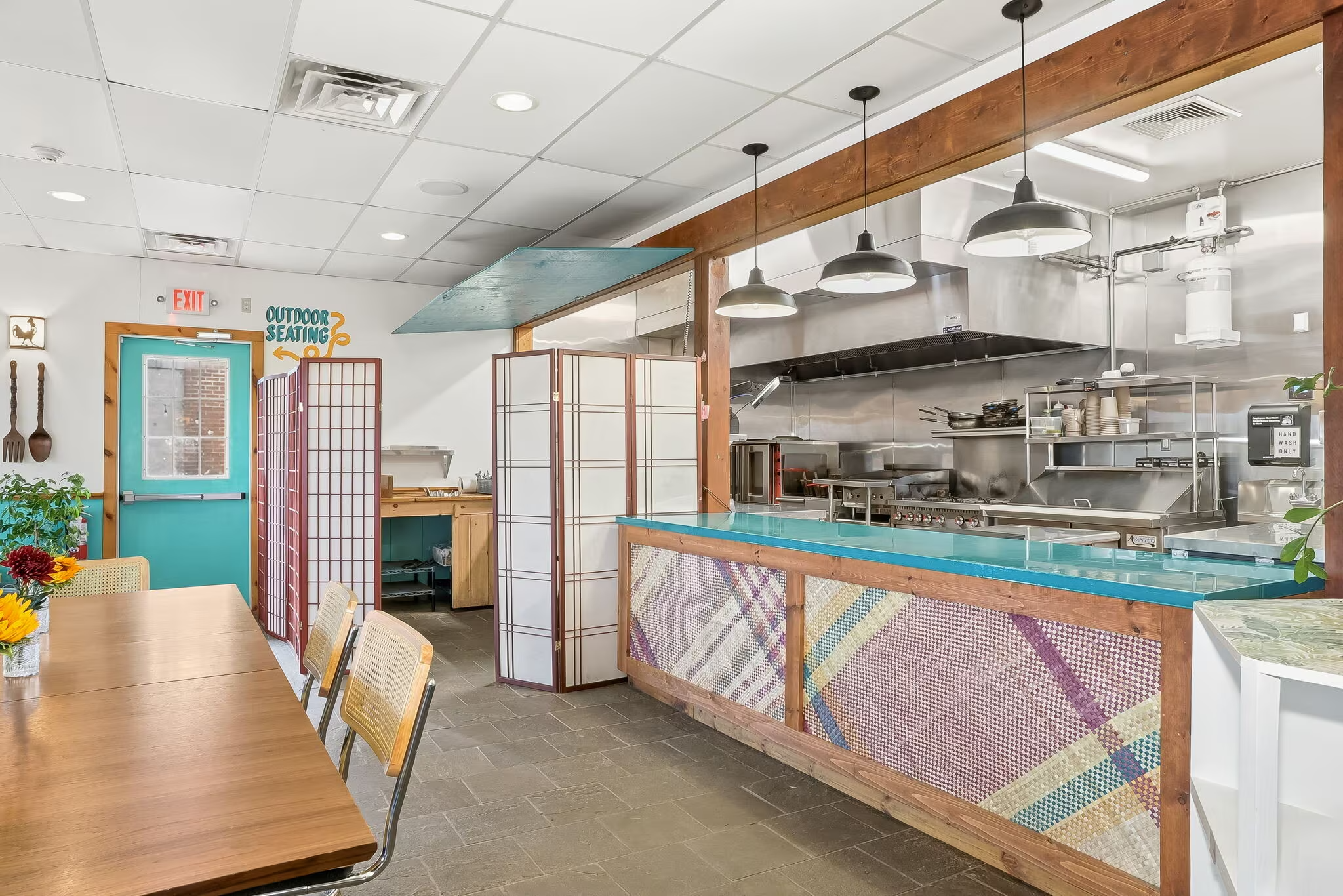Interior of a restaurant showing a table with chairs, a kitchen area with stainless steel appliances, shelves with dishes, hanging lights, and a decorative partition.