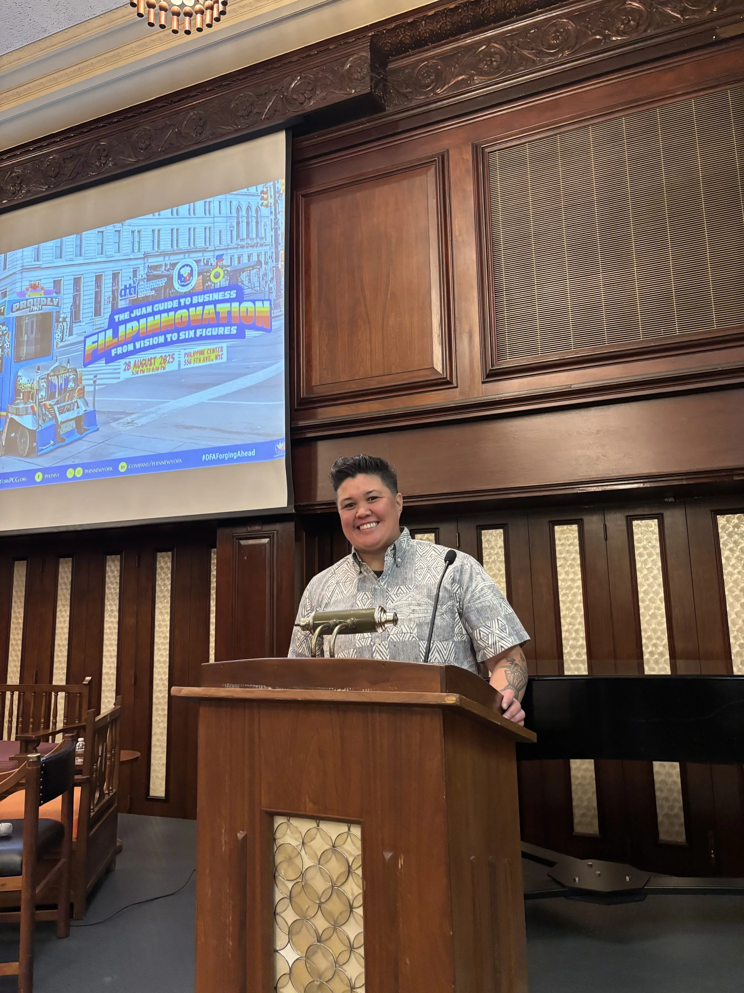 A woman standing at a wooden podium in a room with wood-paneled walls, smiling at the camera, with a microphone attached to the podium. There is a large projection screen behind her displaying a colorful poster about a business event.