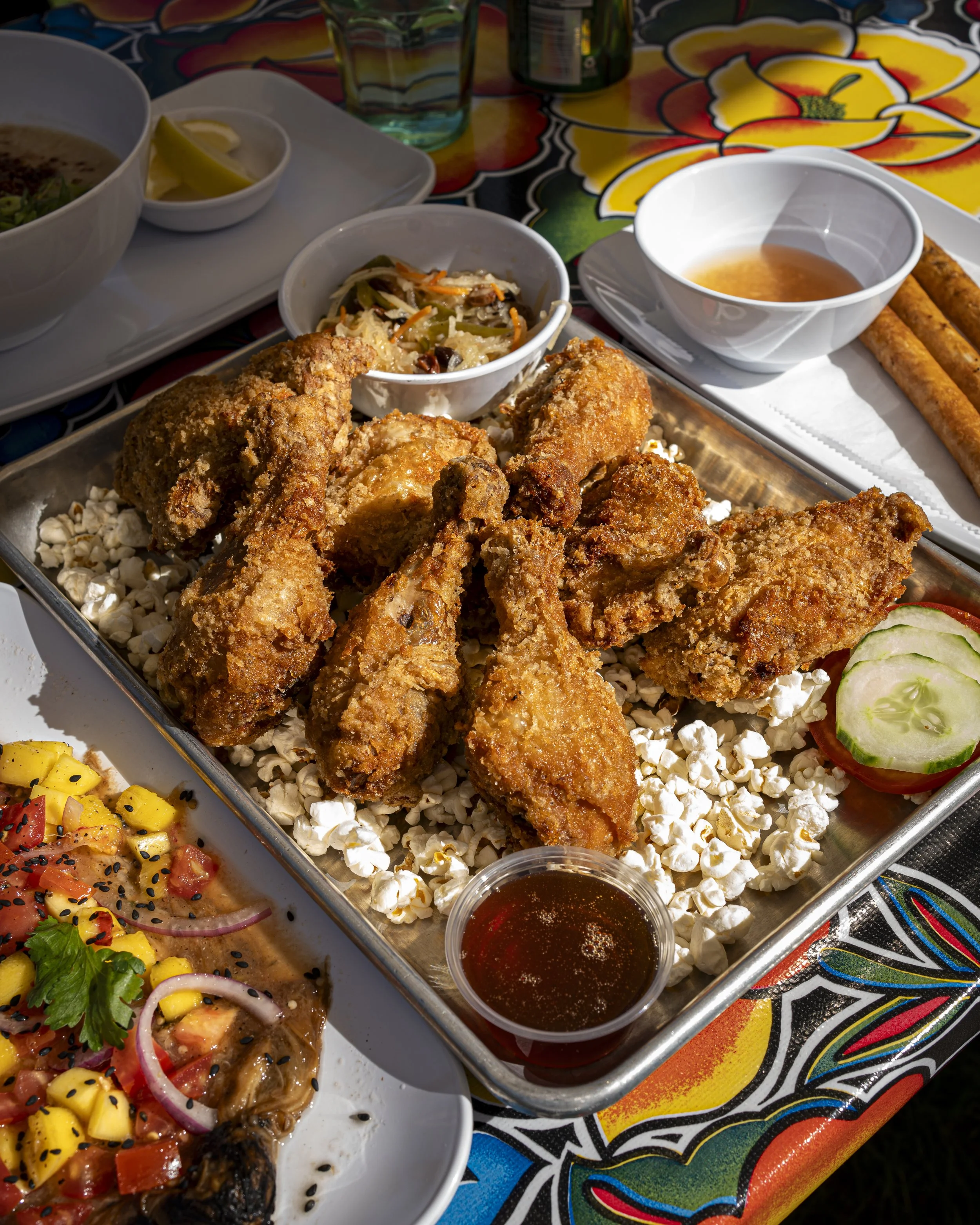 Fried chicken served with popcorn on a tray, with side dishes of small bowl of pickled vegetables, creamy dressing, and chili sauce, on a colorful floral tablecloth.