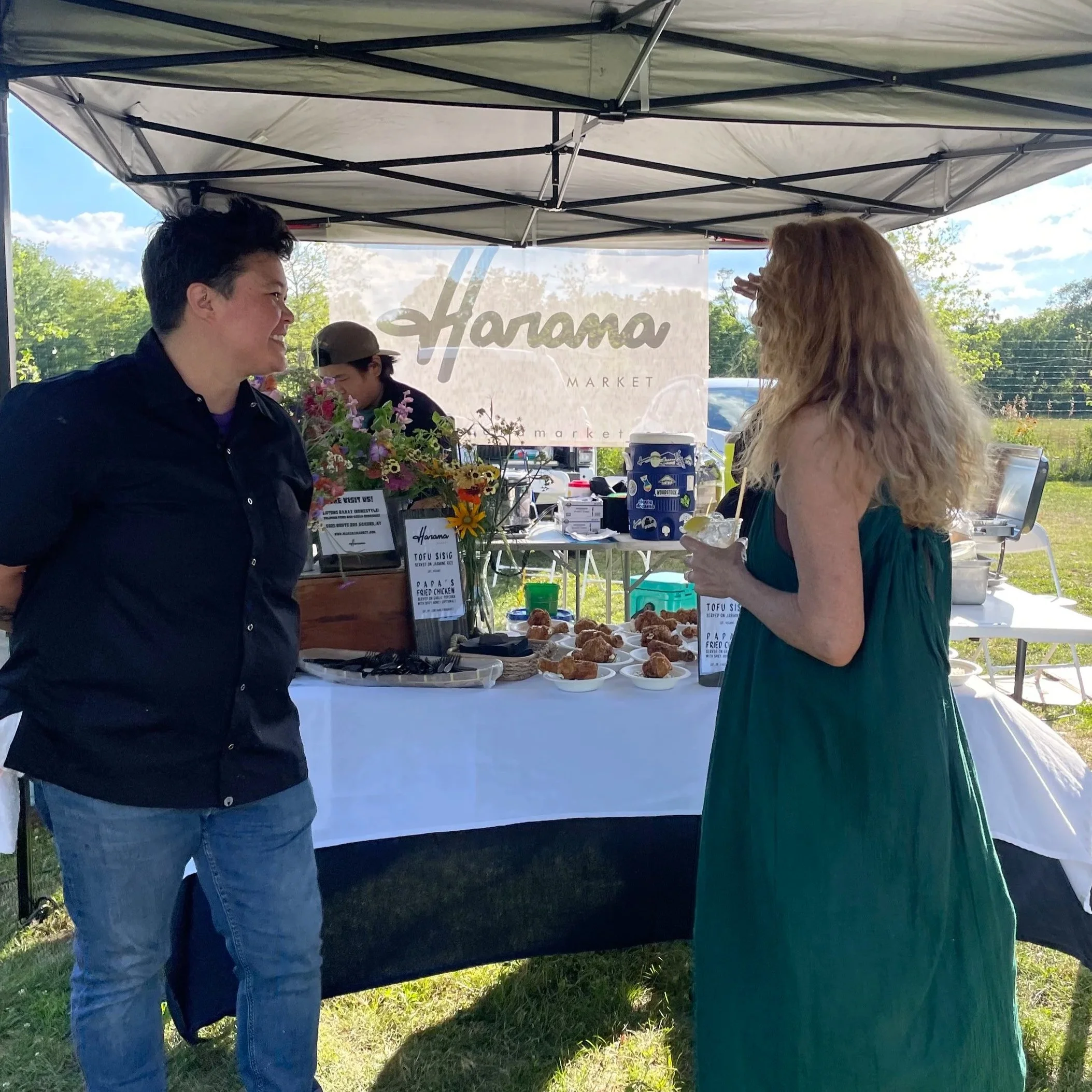 Two women conversing at an outdoor market stand under a canopy, with a table of food and flowers, and a sign that reads 'Harama Market' in the background.