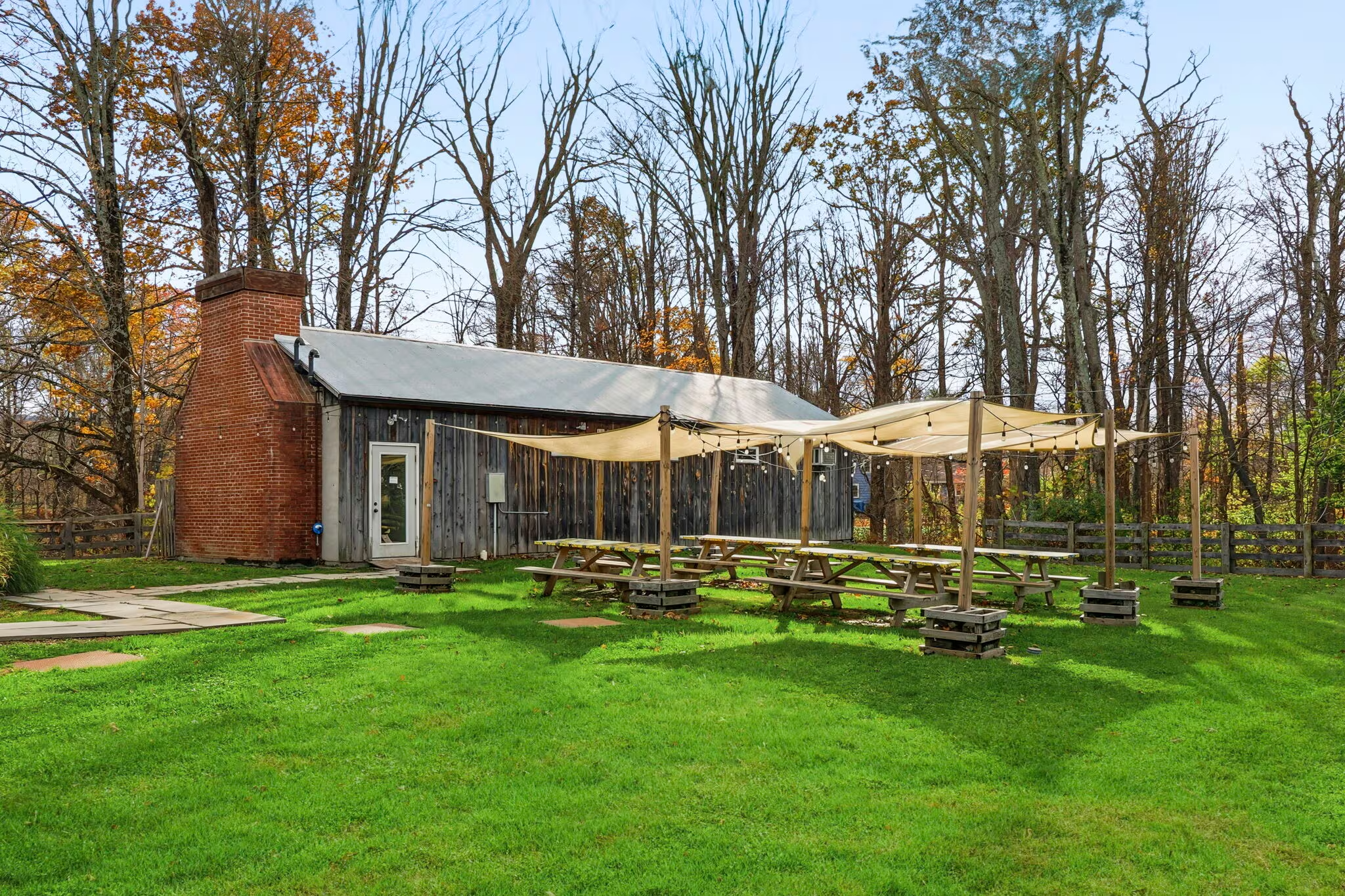 Outdoor scene featuring a rustic building with a brick chimney and wooden siding, surrounded by trees with autumn leaves. There are picnic tables with a canopy and string lights, on a grassy lawn with stone pavers.