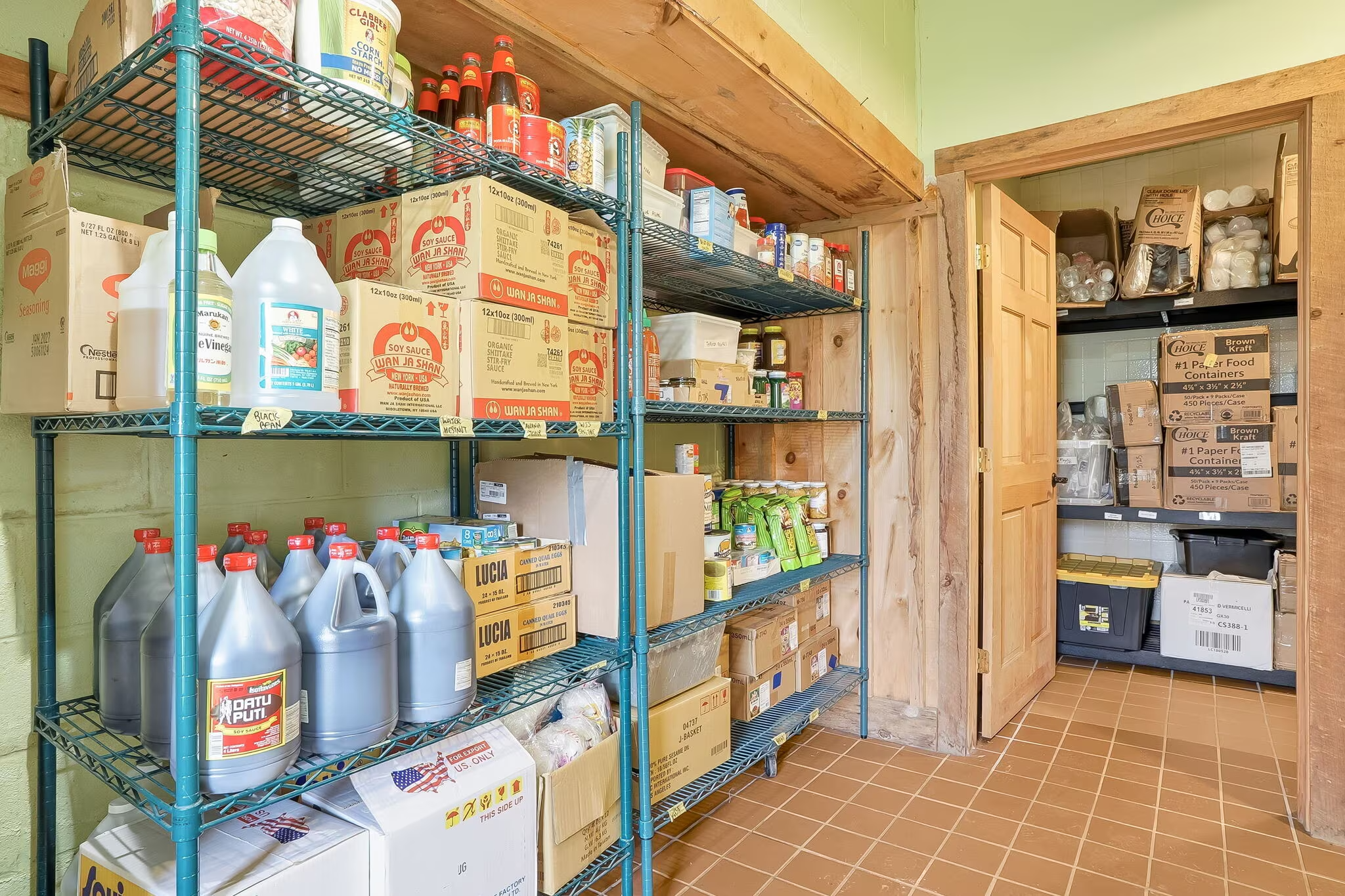 Storage room with shelves stocked with canned goods, bottled sauces, and boxed food items, along with a closet holding paper products and other supplies.