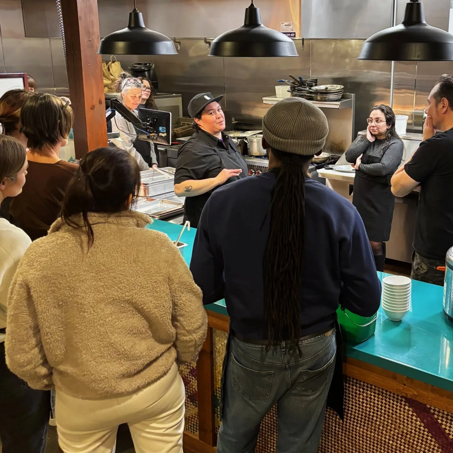 A group of people gathered in a restaurant kitchen listening to a chef explain, with several people wearing aprons and head coverings, and others standing around a counter.