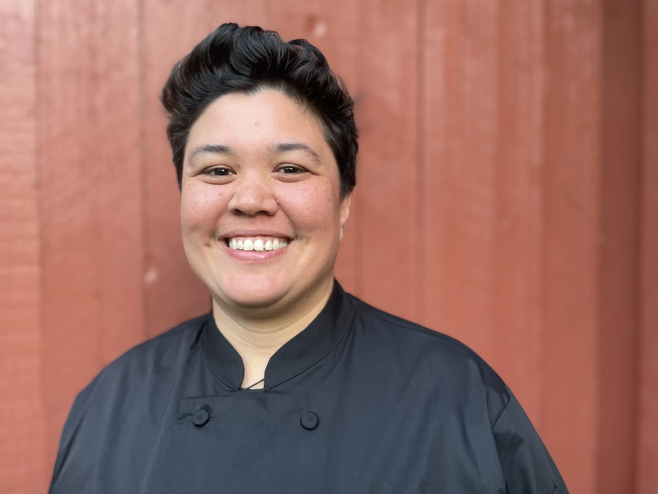 A smiling woman with short dark hair wearing a black chef's coat, standing in front of a reddish-brown wooden wall.