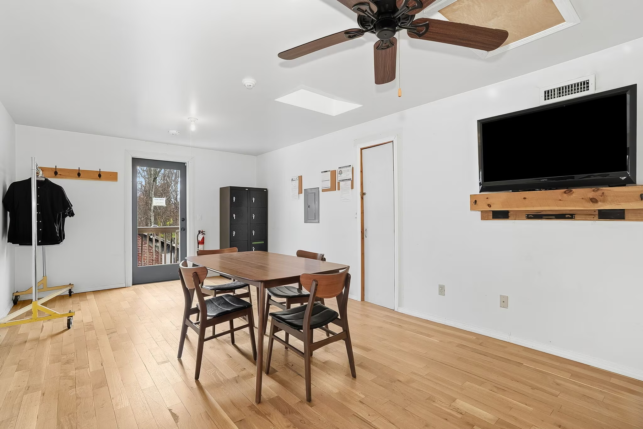 A dining area with a wooden table and four chairs, a ceiling fan, a wall-mounted TV on a wooden shelf, a black storage cabinet, and a door leading outside. The room has white walls and light wood flooring.