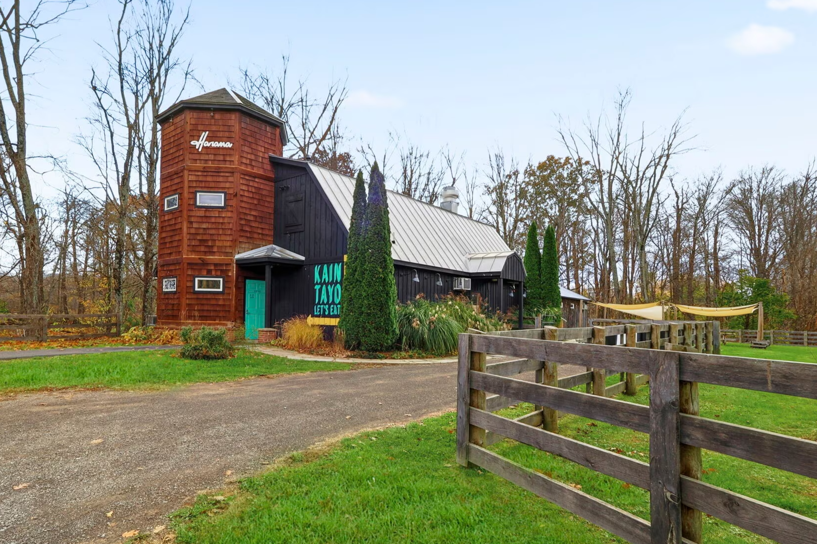 A building with a tower made of reddish wood on the left and black-painted sections on the right, surrounded by green grass, trees, and fenced areas in the background, with a sign that says "KAIM JAYO LET'S EAT" in front.