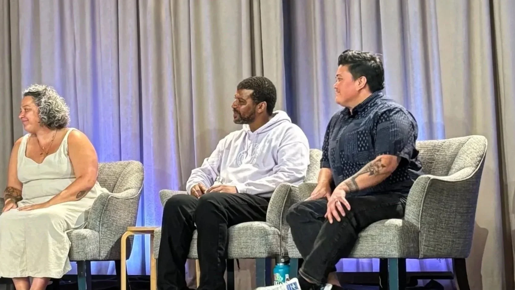 Three people seated on a stage during a panel discussion with grey curtains in the background.