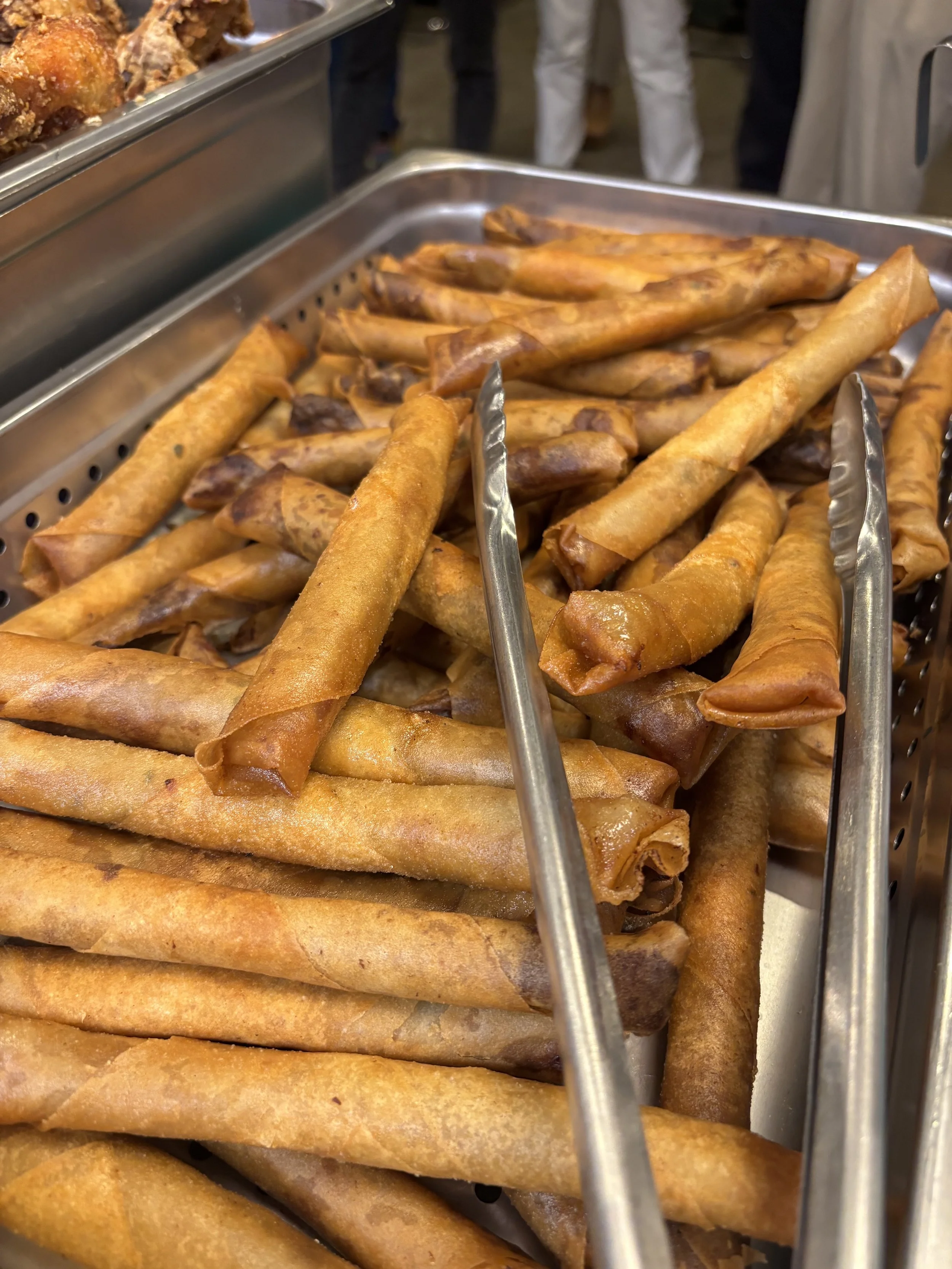 Fried spring rolls in a stainless steel tray, set in a buffet or food service setting.