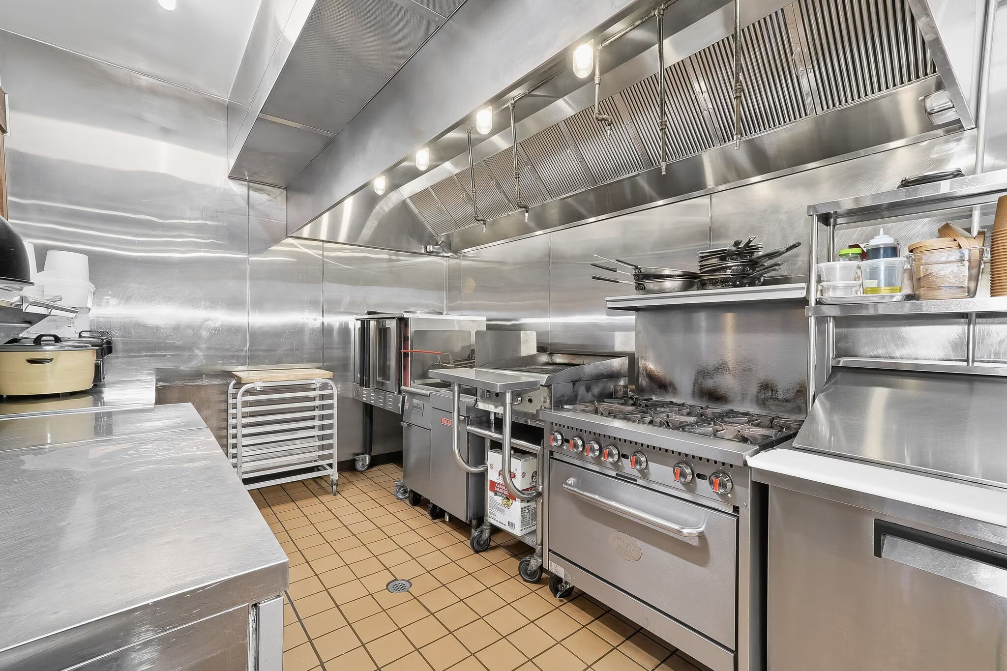 Commercial kitchen with stainless steel appliances, stovetop, oven, microwave, and shelves, tiled floor, and overhead lighting.