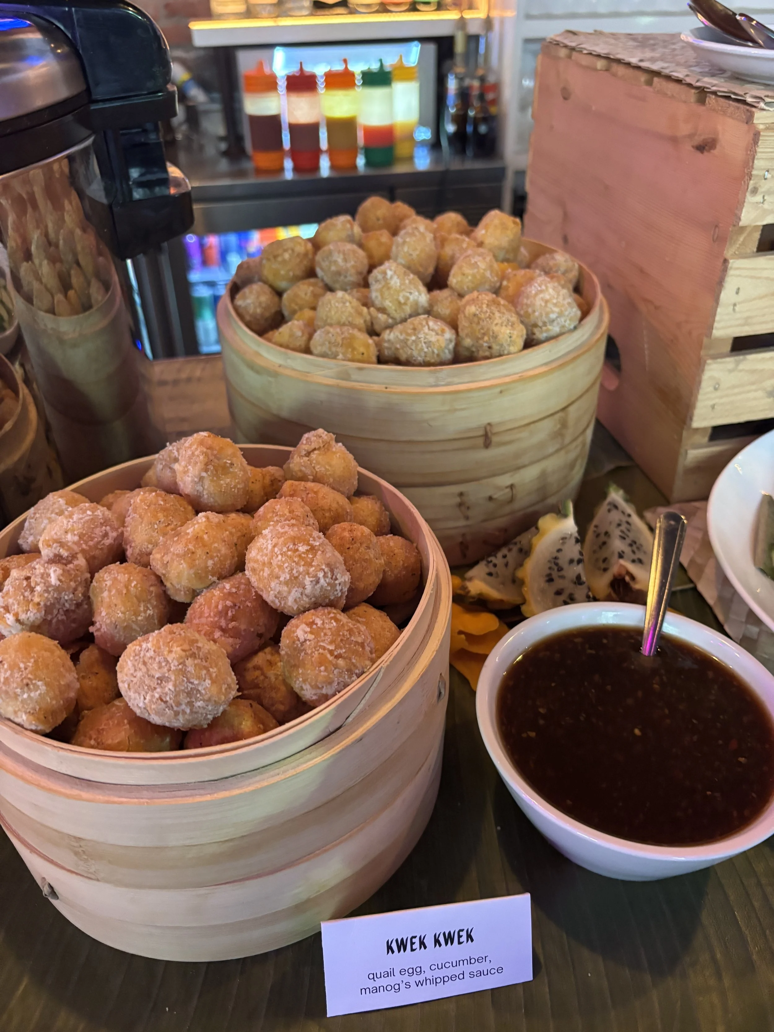 Two baskets of fried quail eggs, cucumber, and mango with a bowl of whipped sauce on a restaurant table with condiment bottles in the background.