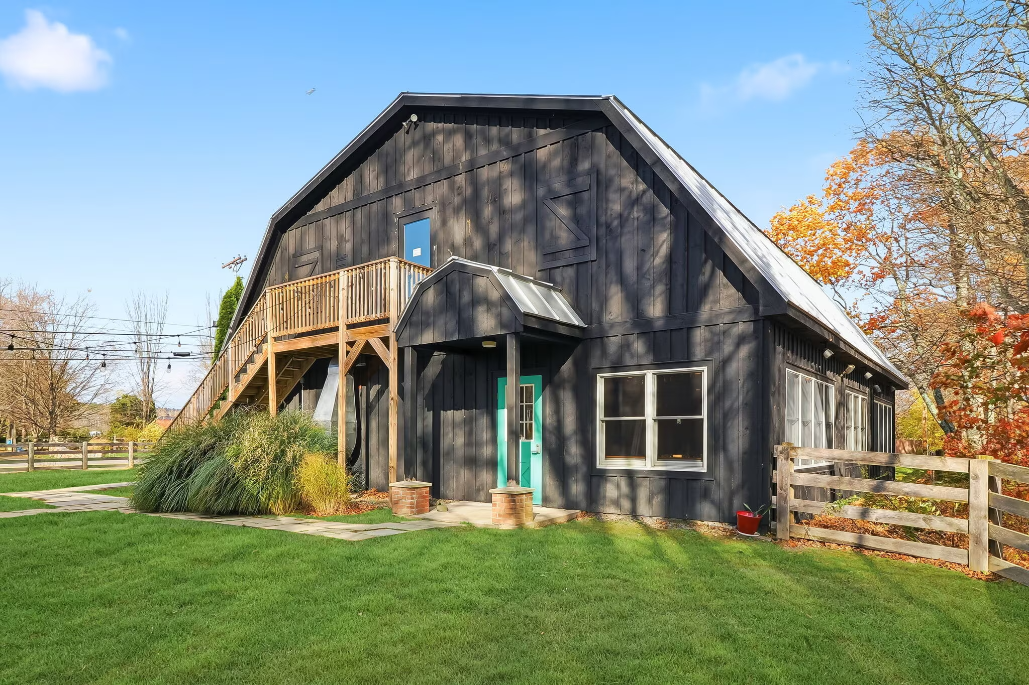 A black barn-style house with a small balcony, a wooden staircase, and a teal front door, surrounded by green grass and trees with autumn leaves.