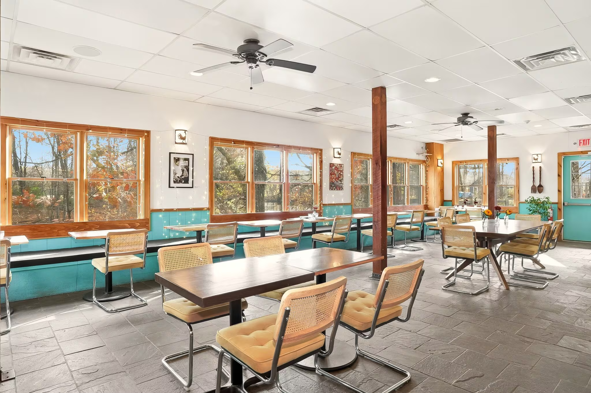 Empty dining area with wooden tables and yellow-cushioned chairs, large windows with outside view, ceiling fans, and decorative wall art.