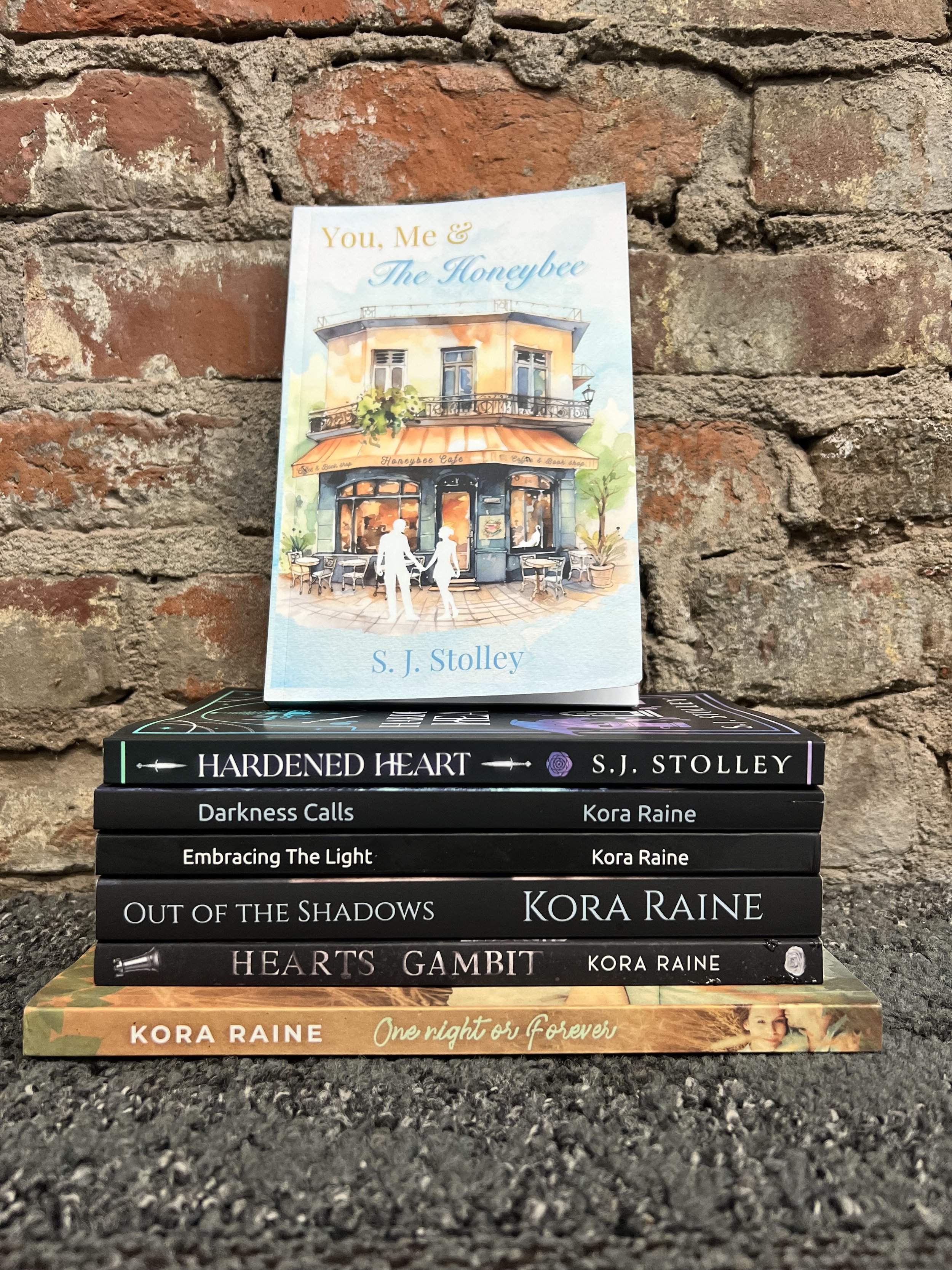 Stack of six books against a brick wall, with a watercolor-style book titled "You, Me & The Honeybee" on top, depicting a cafe scene with an outdoor patio and two white silhouettes of a couple holding hands.