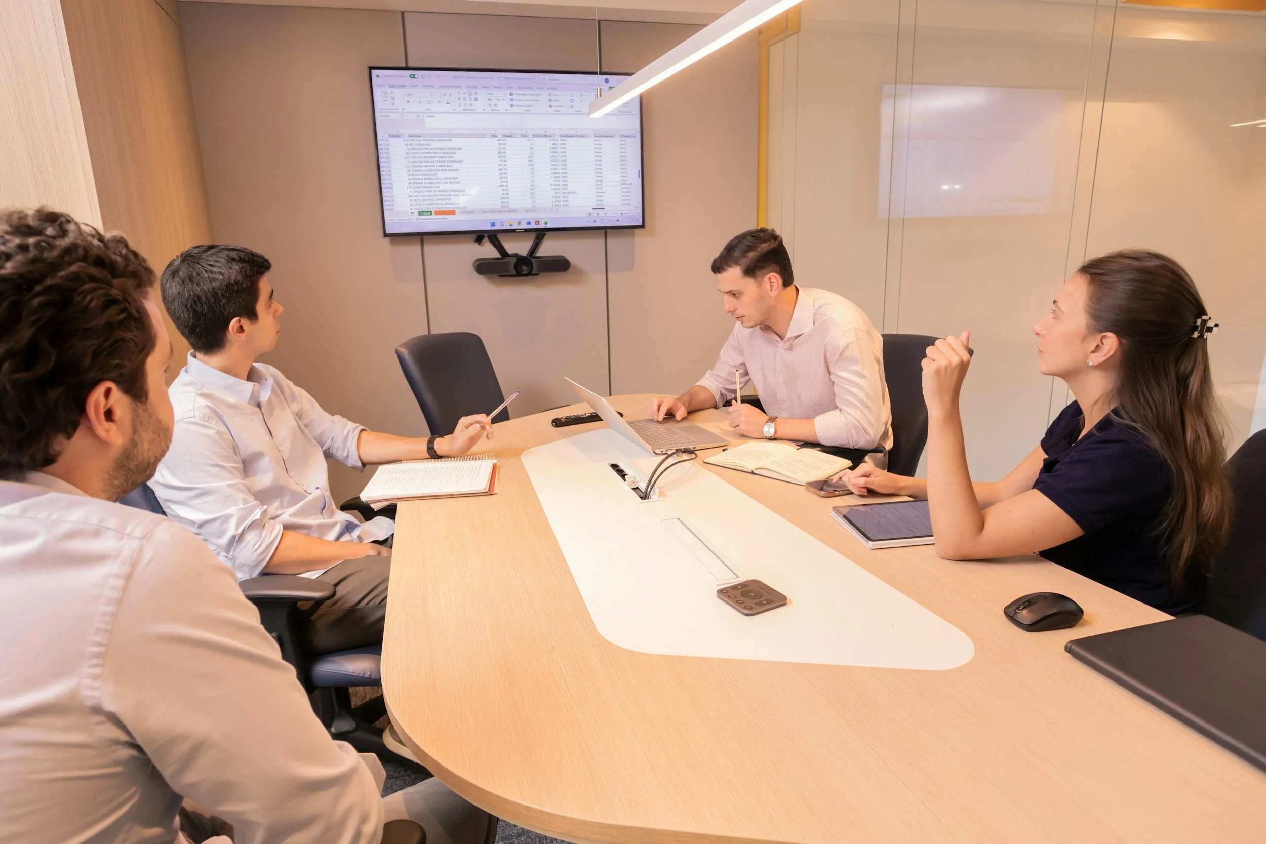 A business meeting with five people around a conference table, two women and three men, engaged in discussion, with a large monitor displaying a spreadsheet on the wall.