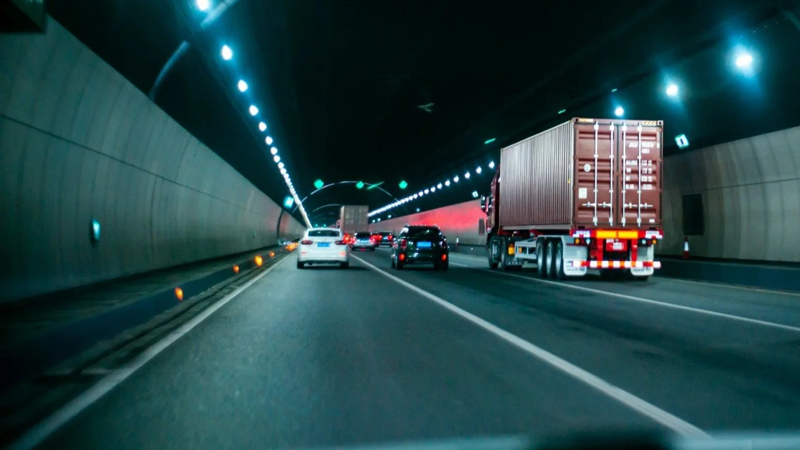 Vehicles driving through a tunnel, including a large red truck, cars, and a white vehicle, with illuminated lights on the ceiling and walls.