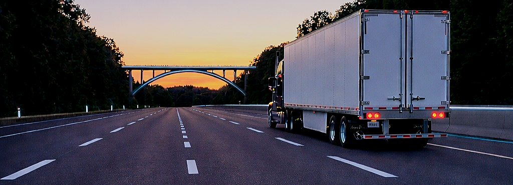 A semi-truck driving on an empty highway at sunset, with a bridge and trees in the background.