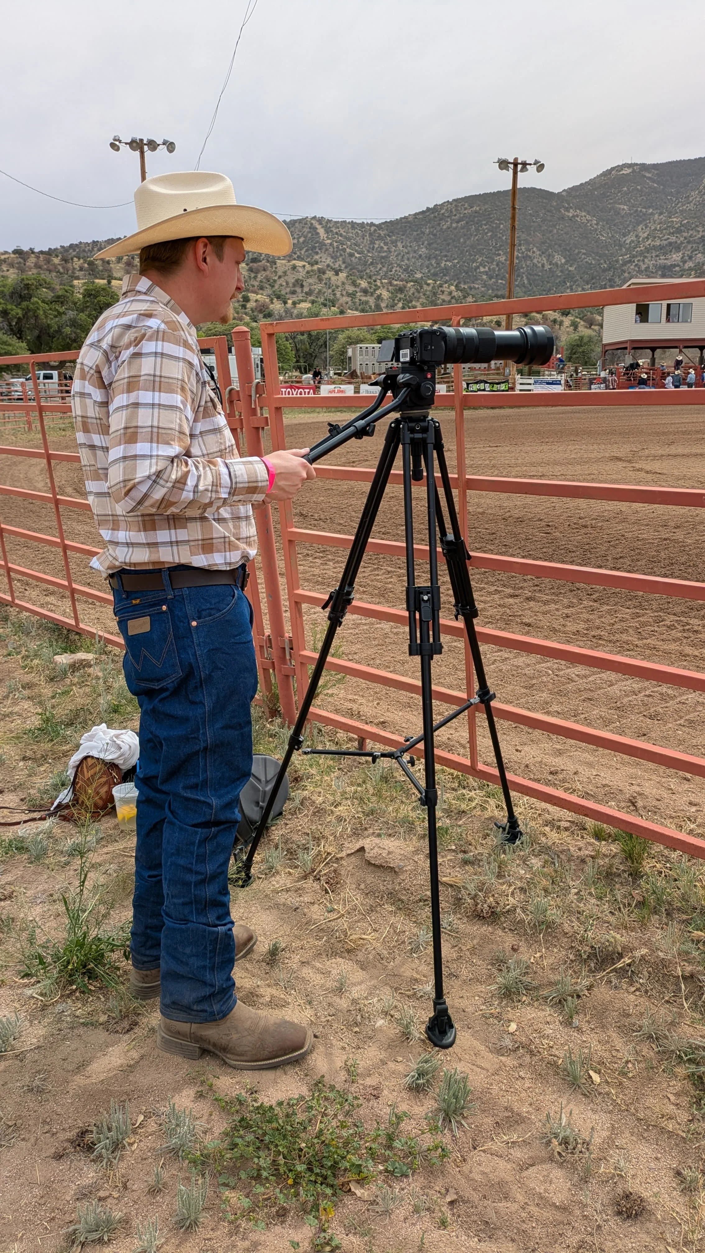 A person wearing a cowboy hat and plaid shirt standing on dirt ground behind a pink metal fence, operating a professional camera on a tripod at an outdoor rodeo or similar event, with hills and utility poles in the background.