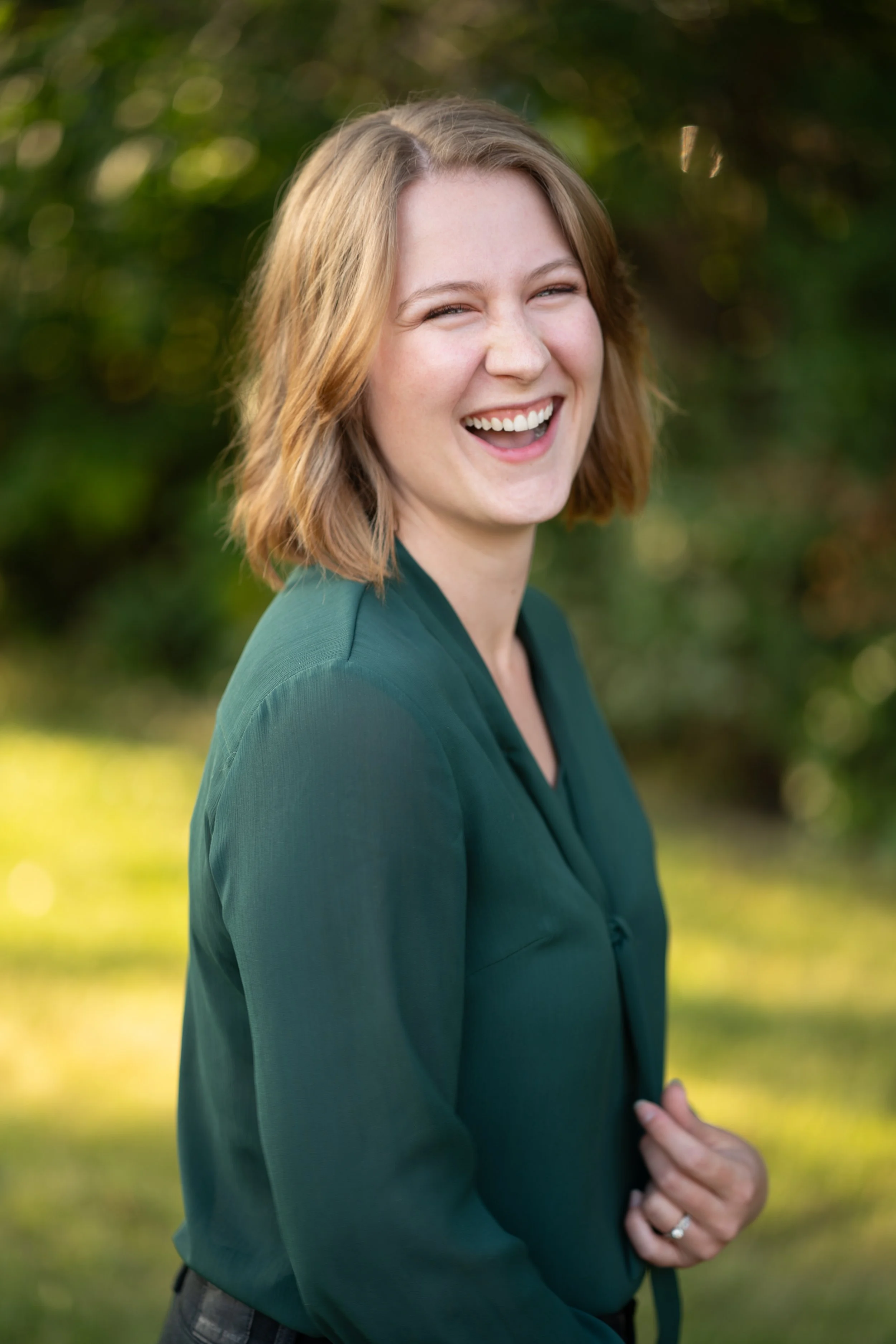 A woman with short, light brown hair is smiling and looking at the camera. She is outside with a blurry background of green trees and sunlight.