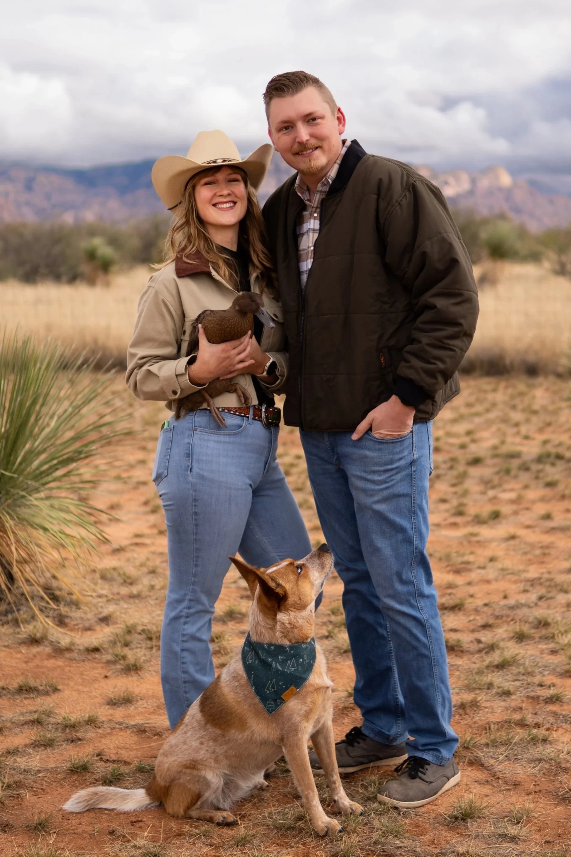 A smiling couple with a dog and a duck in a desert landscape with mountains in the background.