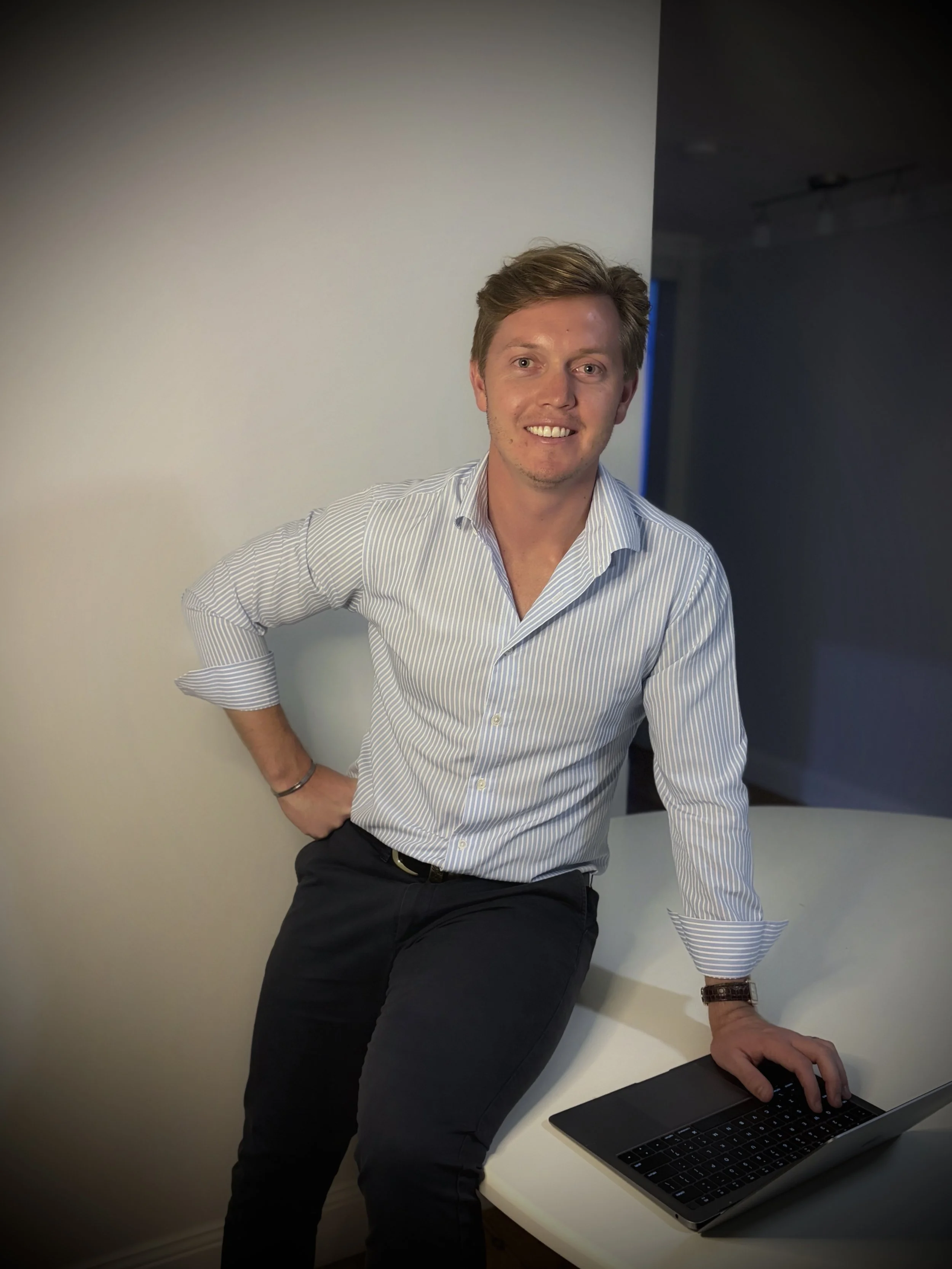 A man in a striped shirt and dark pants sitting at a white desk with a laptop, in an office setting.