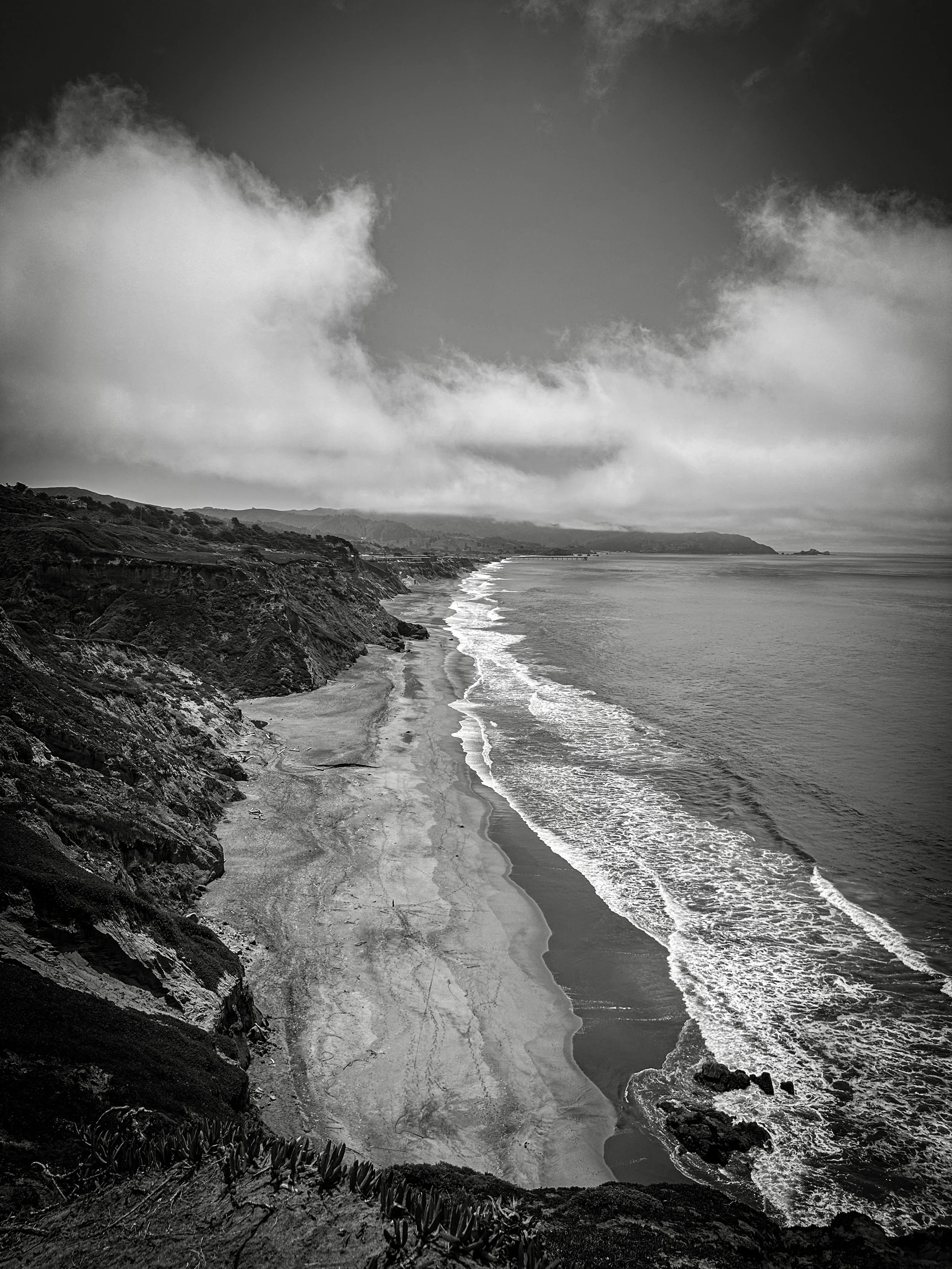 A black and white photo of a rugged coastline with steep cliffs and a sandy beach beneath. Ocean waves are crashing along the shore, and clouds are scattered across the sky.