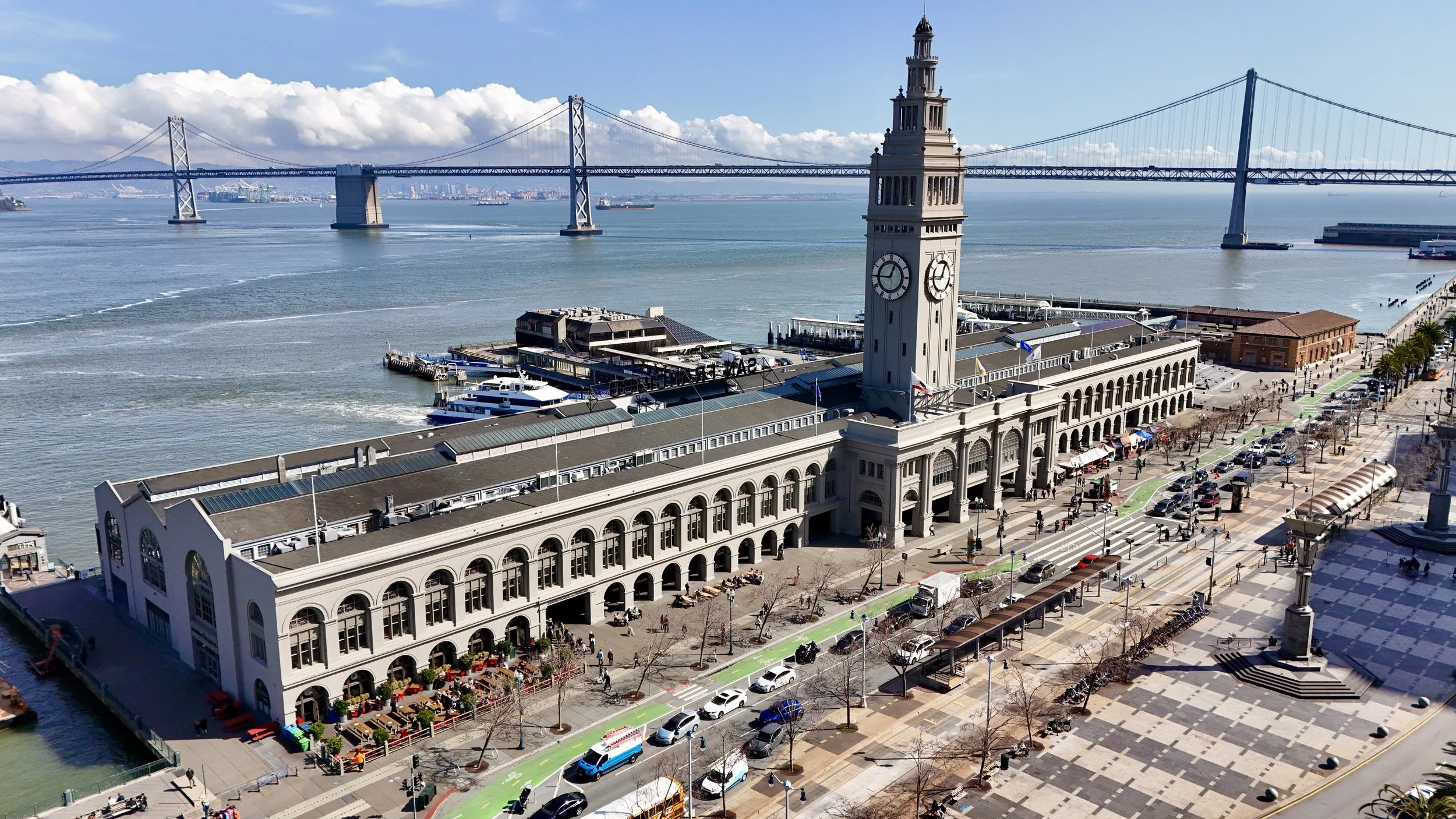 Aerial view of San Francisco Ferry Building with a clock tower, waterfront, and Bay Bridge in the background, with boats in the bay, cars and pedestrians on the street, and market stalls outside.