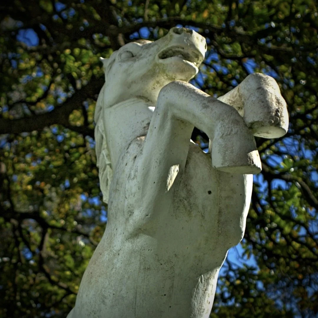 A close-up view of a white horse statue with a raised front leg, set against a background of green leaves and a blue sky.