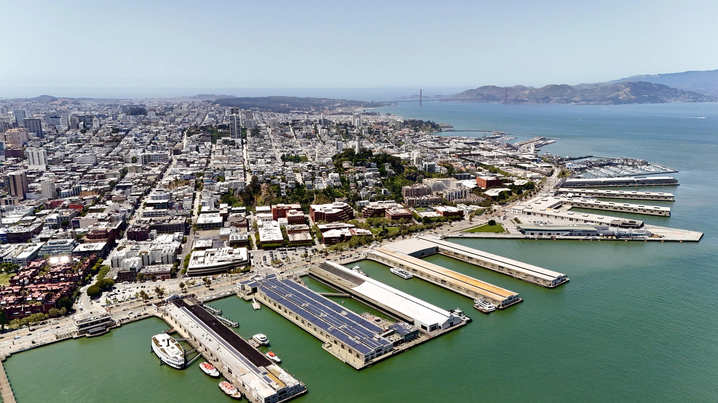 Aerial view of San Francisco waterfront with piers, boats, and the city skyline with hills in the background.