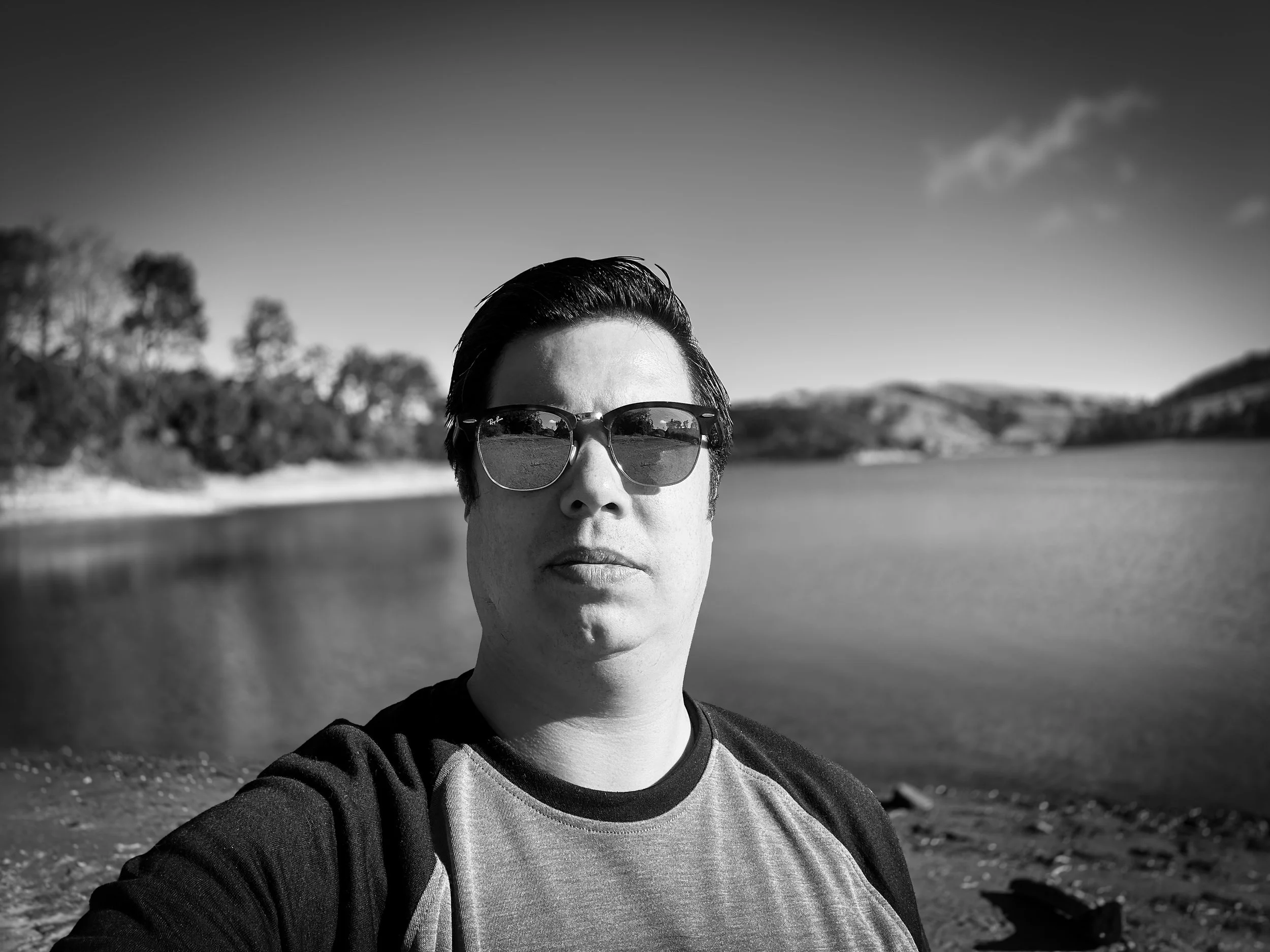 A person wearing sunglasses taking a selfie near a body of water with hills in the background, in black and white.