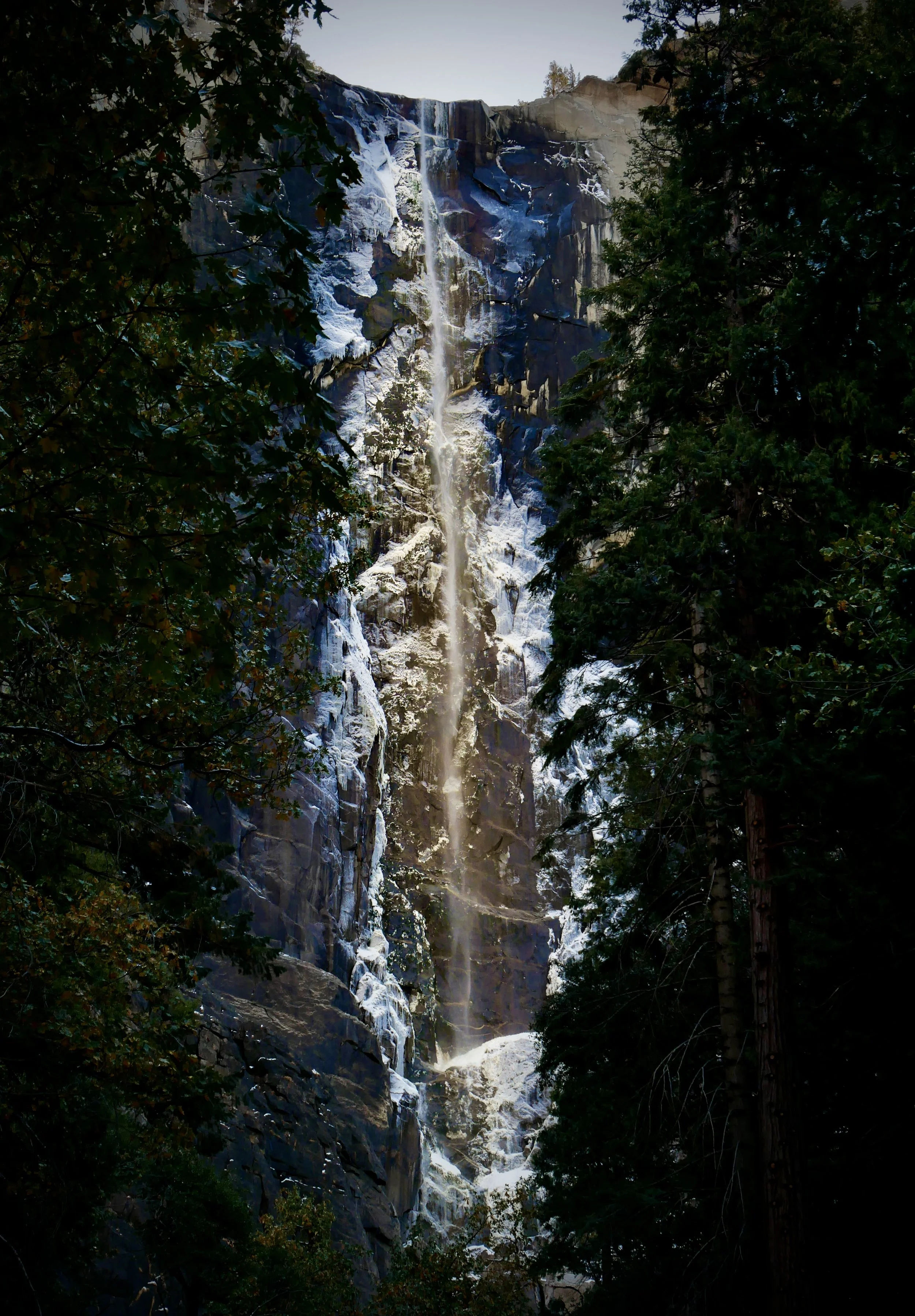 A tall, thin waterfall cascading down a steep, rocky cliff surrounded by trees with snow or ice on the rocks.