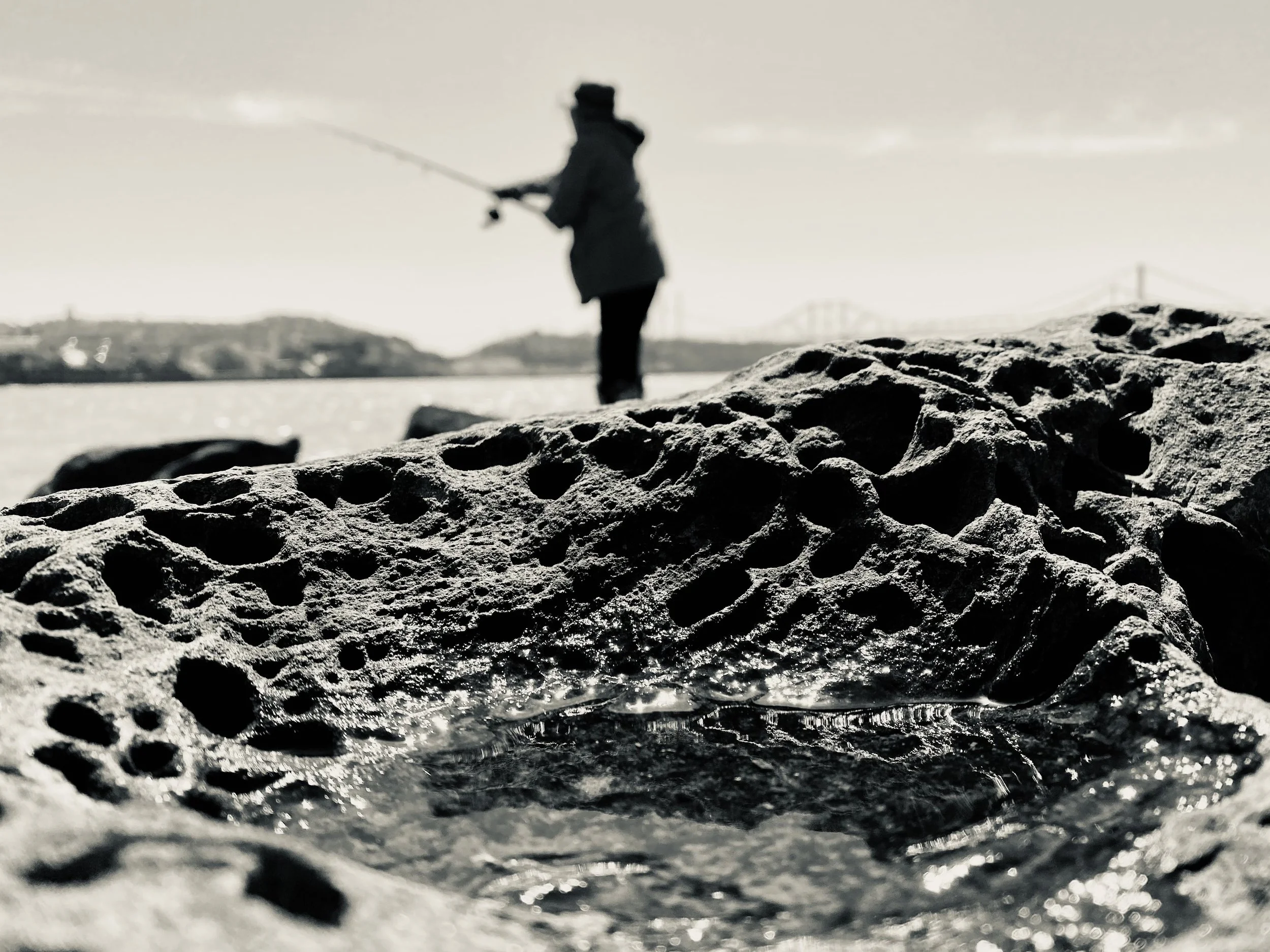 Close-up of a textured rock with a small pool of water, with a person fishing in the background near the riverbank and a bridge in the distance.